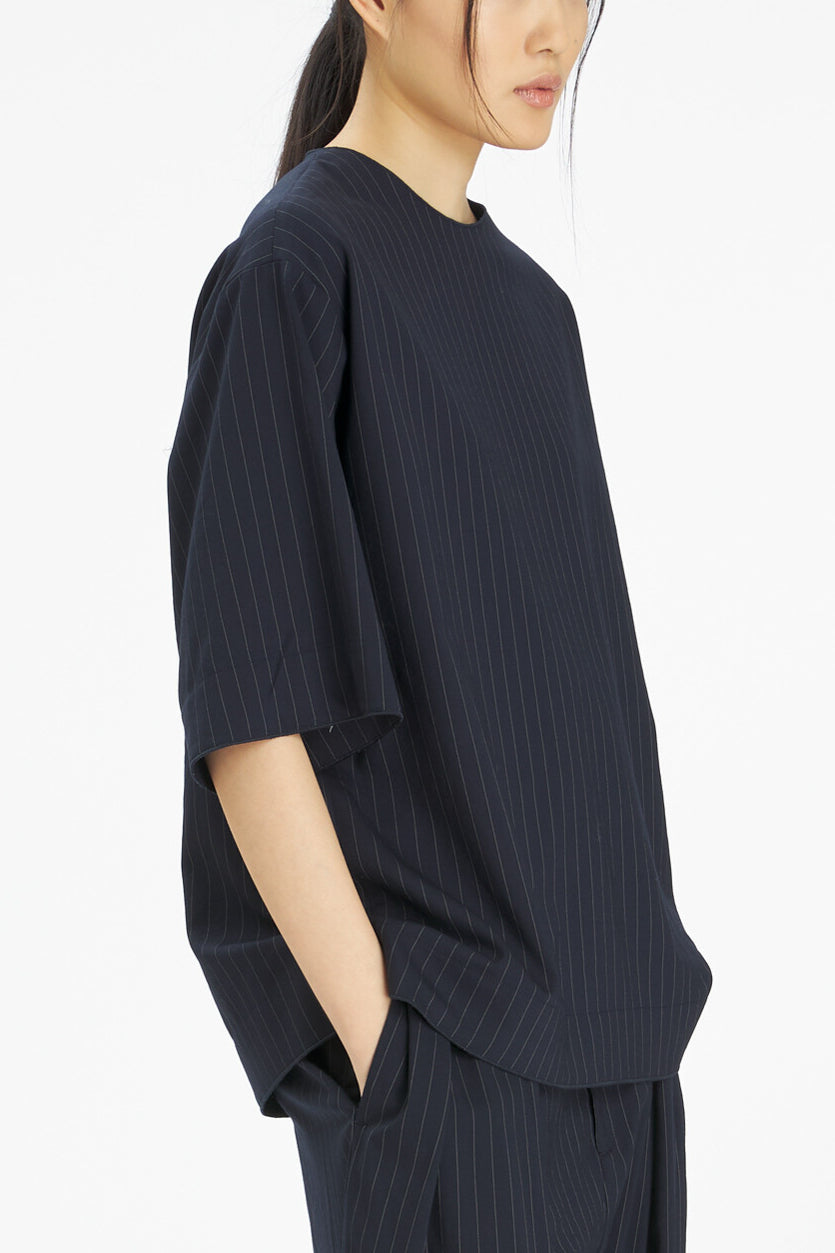 A model shown from the side wearing an oversized dark navy short-sleeve top with thin vertical pinstripes, paired with matching pinstriped trousers, against a clean white background.
