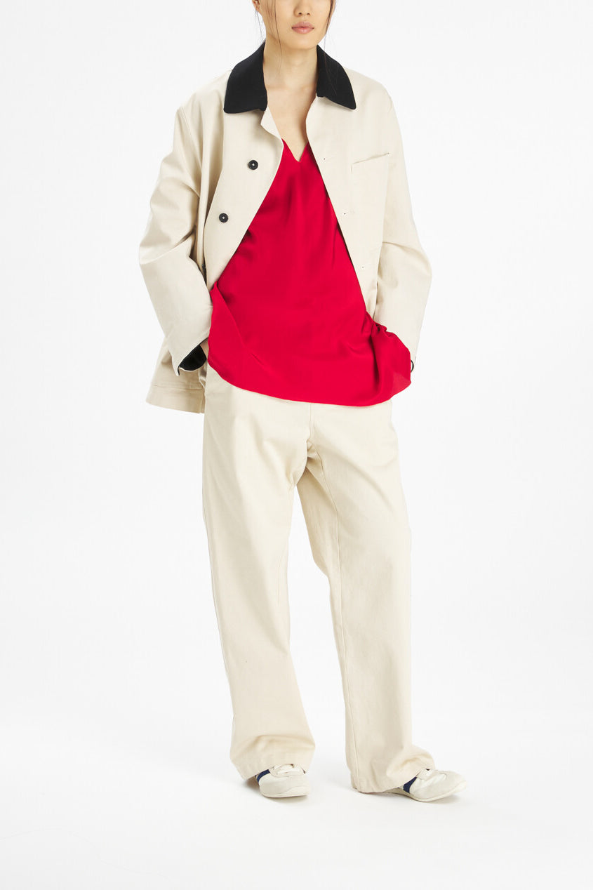 A model wearing a cream-colored jacket with a black collar, matching cream trousers, and a bright red top, standing against a plain white studio background.