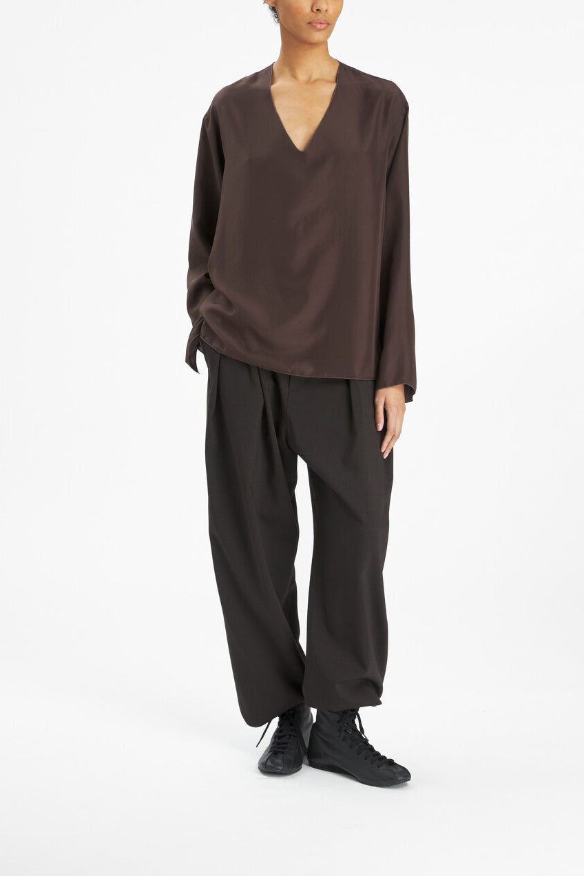 A model wearing a dark brown long-sleeve V-neck top, loose black pants, and black sneakers, photographed against a seamless white studio background.