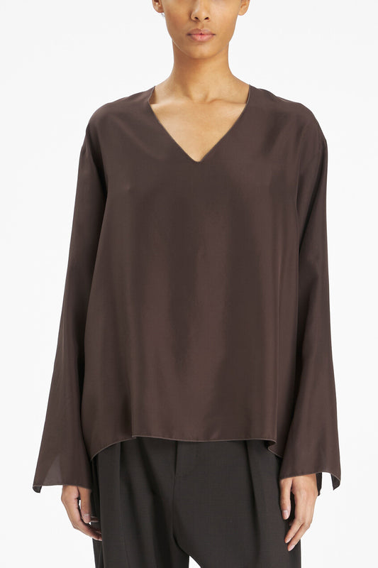 A model wearing a dark brown, long-sleeved V-neck top with a draped, flowing fabric, paired with dark trousers, against a plain white studio background.