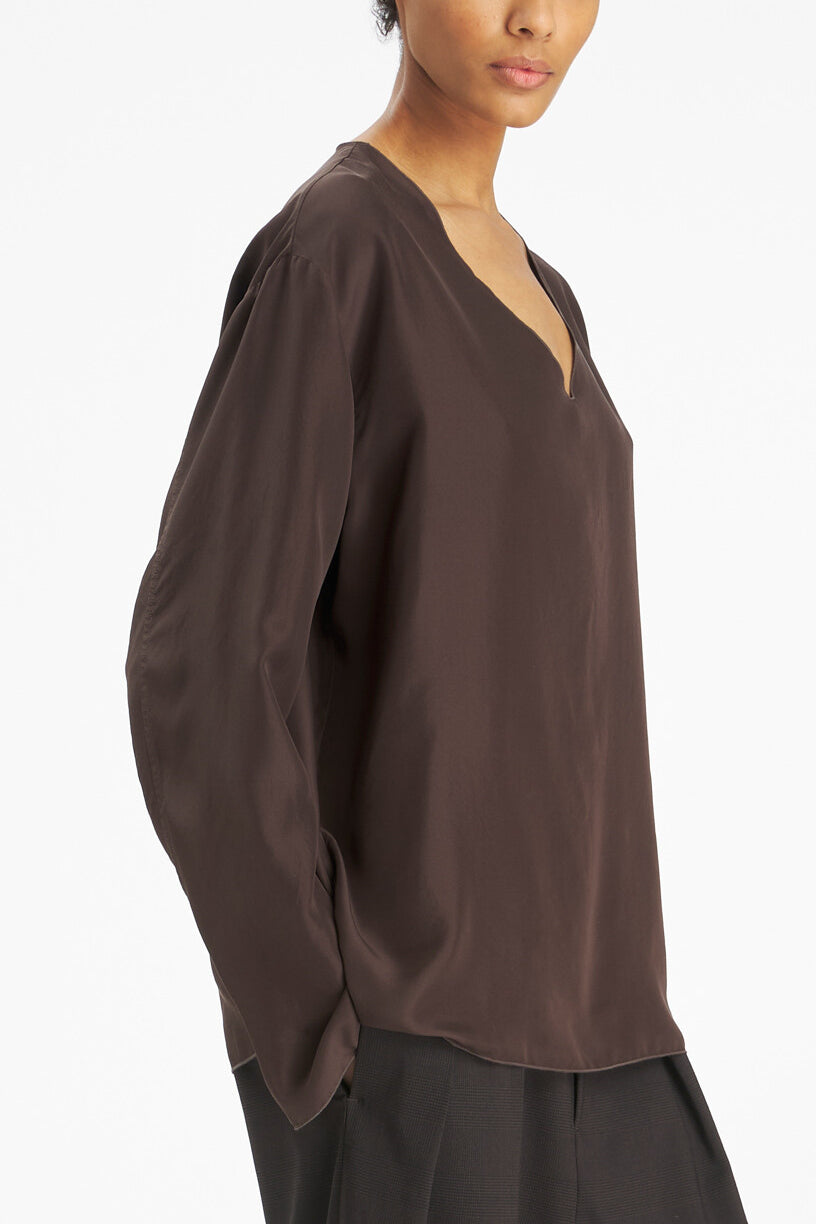 A close-up side view of a model wearing a dark brown, loose-fitting long-sleeve top with a split V-neckline, paired with dark trousers against a plain white background.