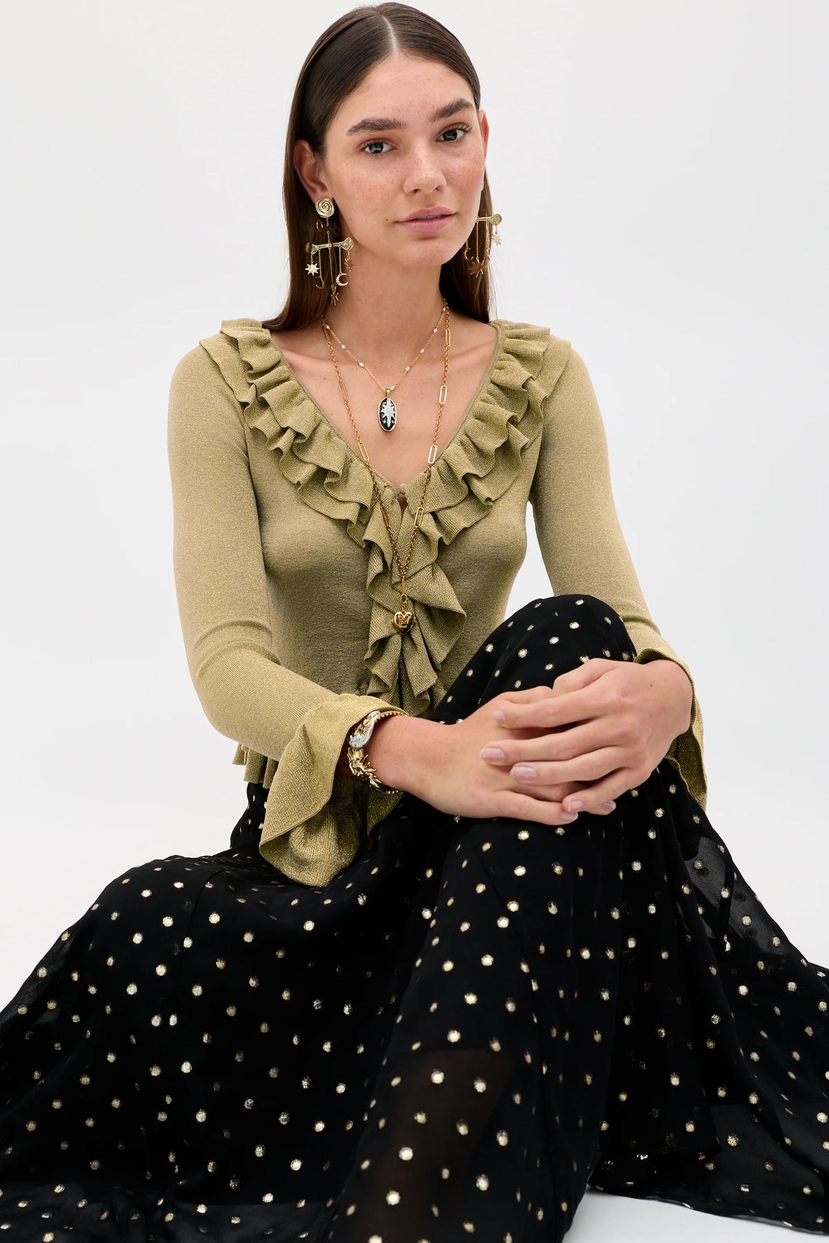 A model wearing a long black skirt with a gold polka-dot pattern, paired with a beige ruffled top and black strappy heels, holding a small black handbag