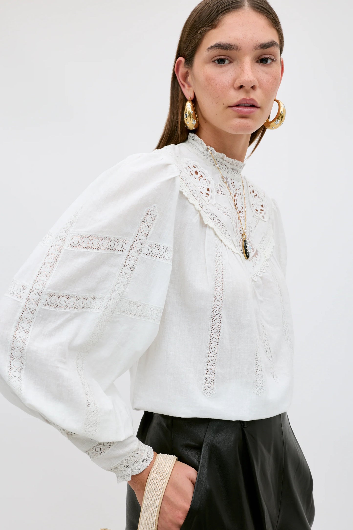 A woman wearing a white long-sleeve blouse with intricate lace and embroidery details, paired with black leather pants, against a plain light gray background.