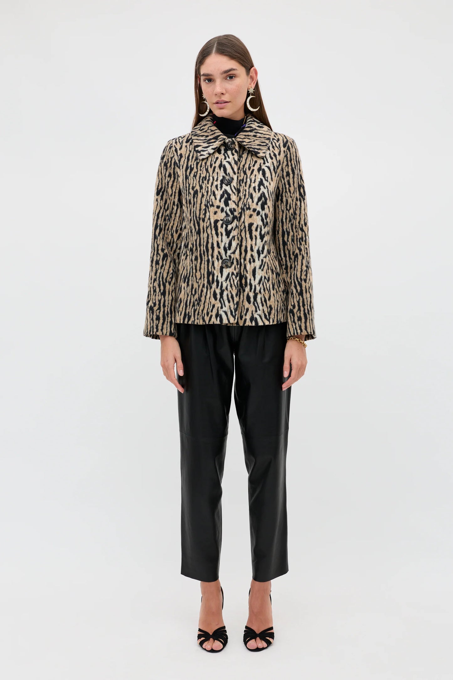A model stands against a plain white studio background wearing a beige and black animal-print jacket with a high collar, black trousers, and black heeled sandals.