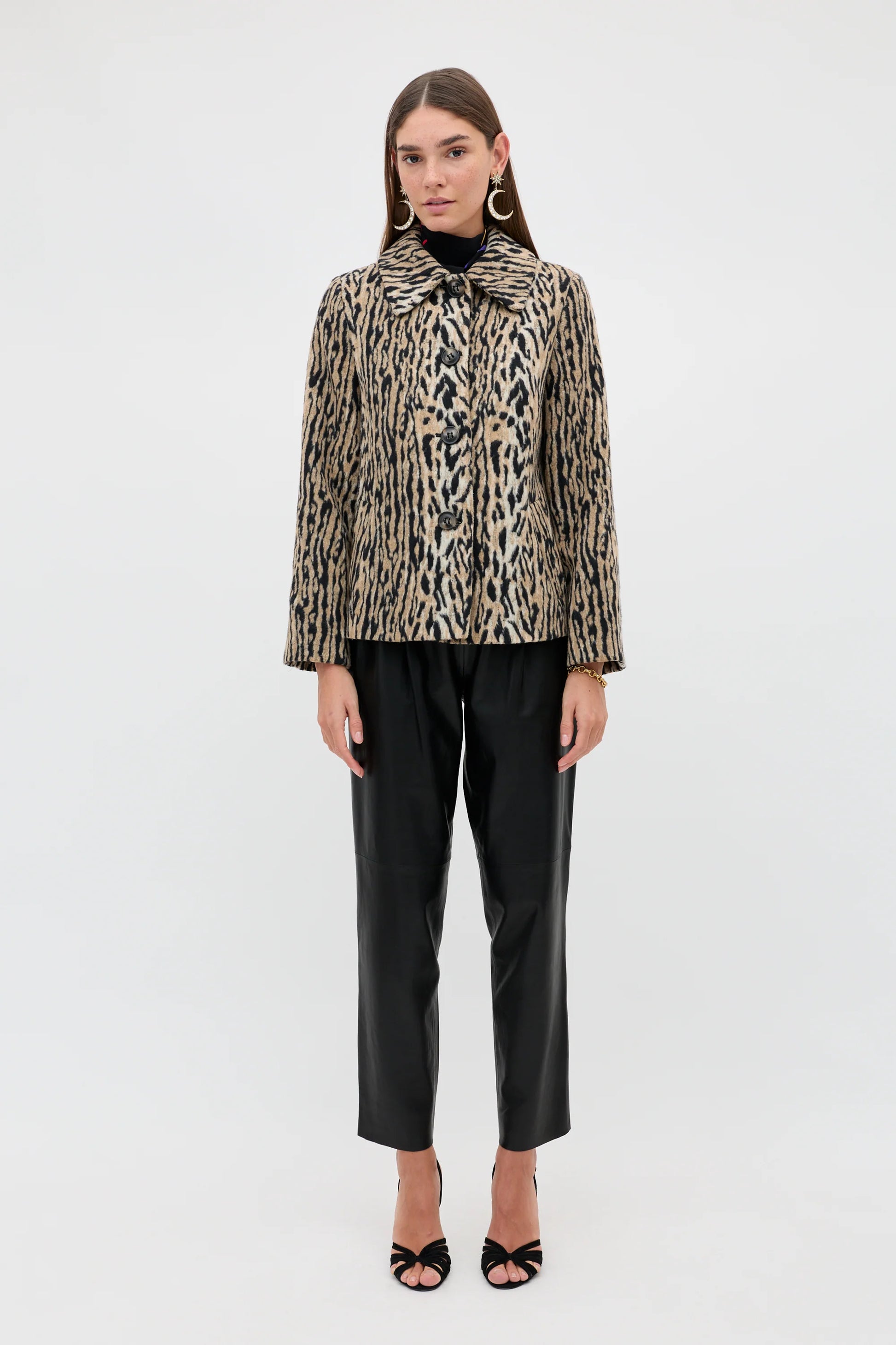 A model stands against a plain white studio background wearing a beige and black animal-print jacket with a high collar, black trousers, and black heeled sandals.