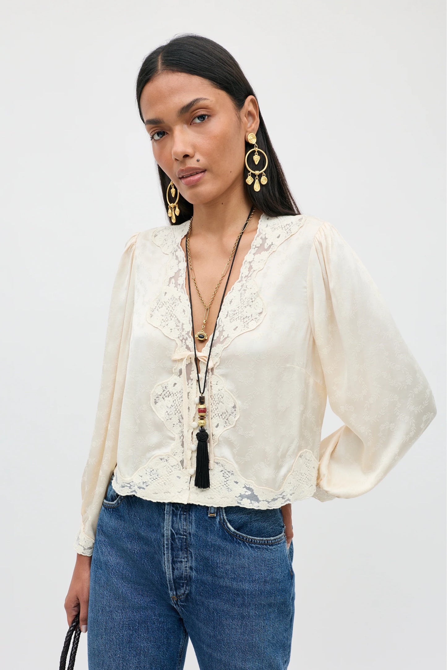 A woman modeling a cream-colored long-sleeve blouse with intricate lace detailing along the V-neck and front, paired with blue jeans and gold jewelry against a plain light gray background.