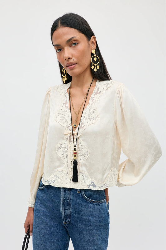 A woman modeling a cream-colored long-sleeve blouse with intricate lace detailing along the V-neck and front, paired with blue jeans and gold jewelry against a plain light gray background.