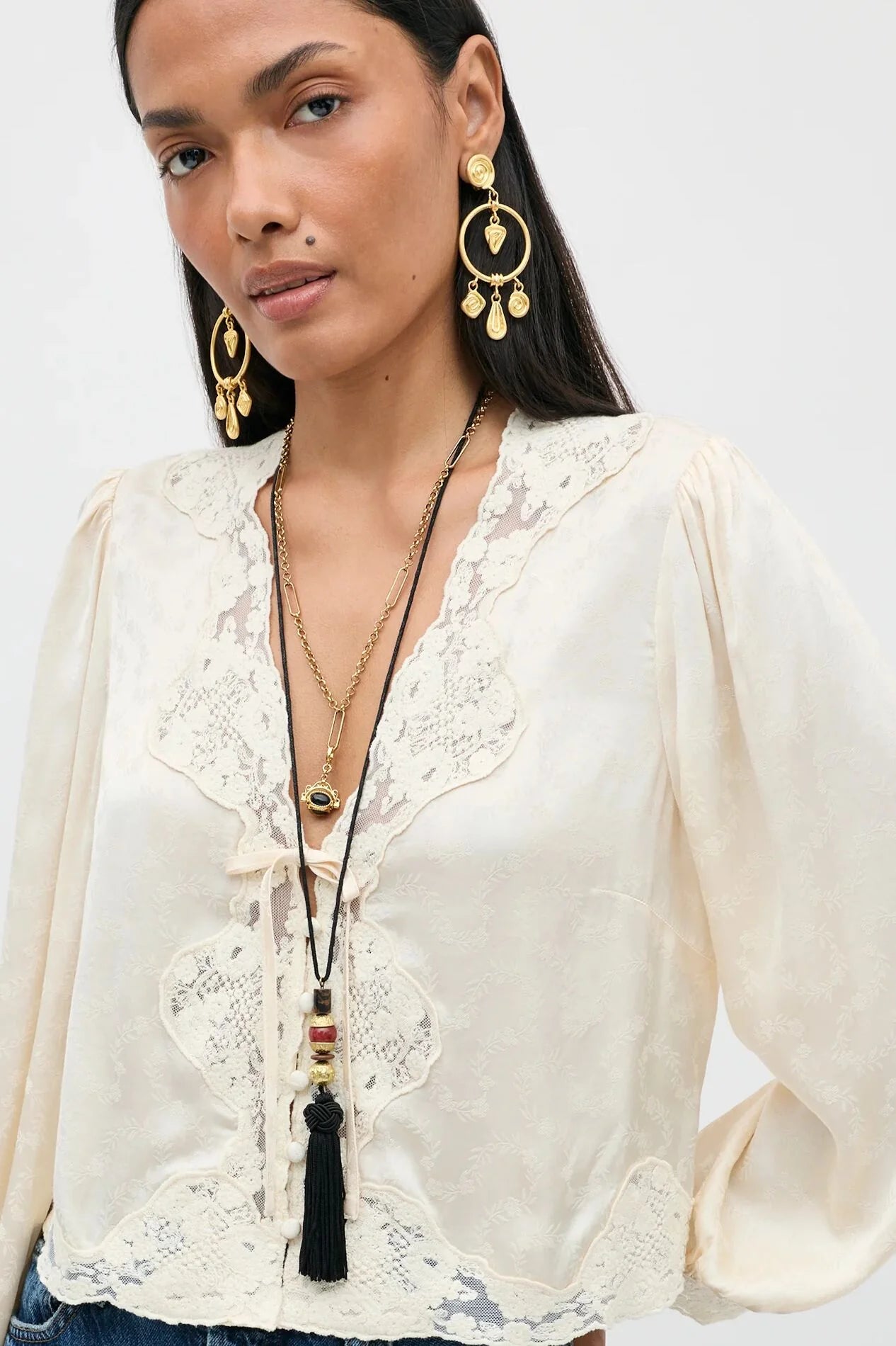 A woman wearing a cream-colored blouse with intricate lace detailing and long balloon sleeves, styled with gold jewelry against a plain light background.