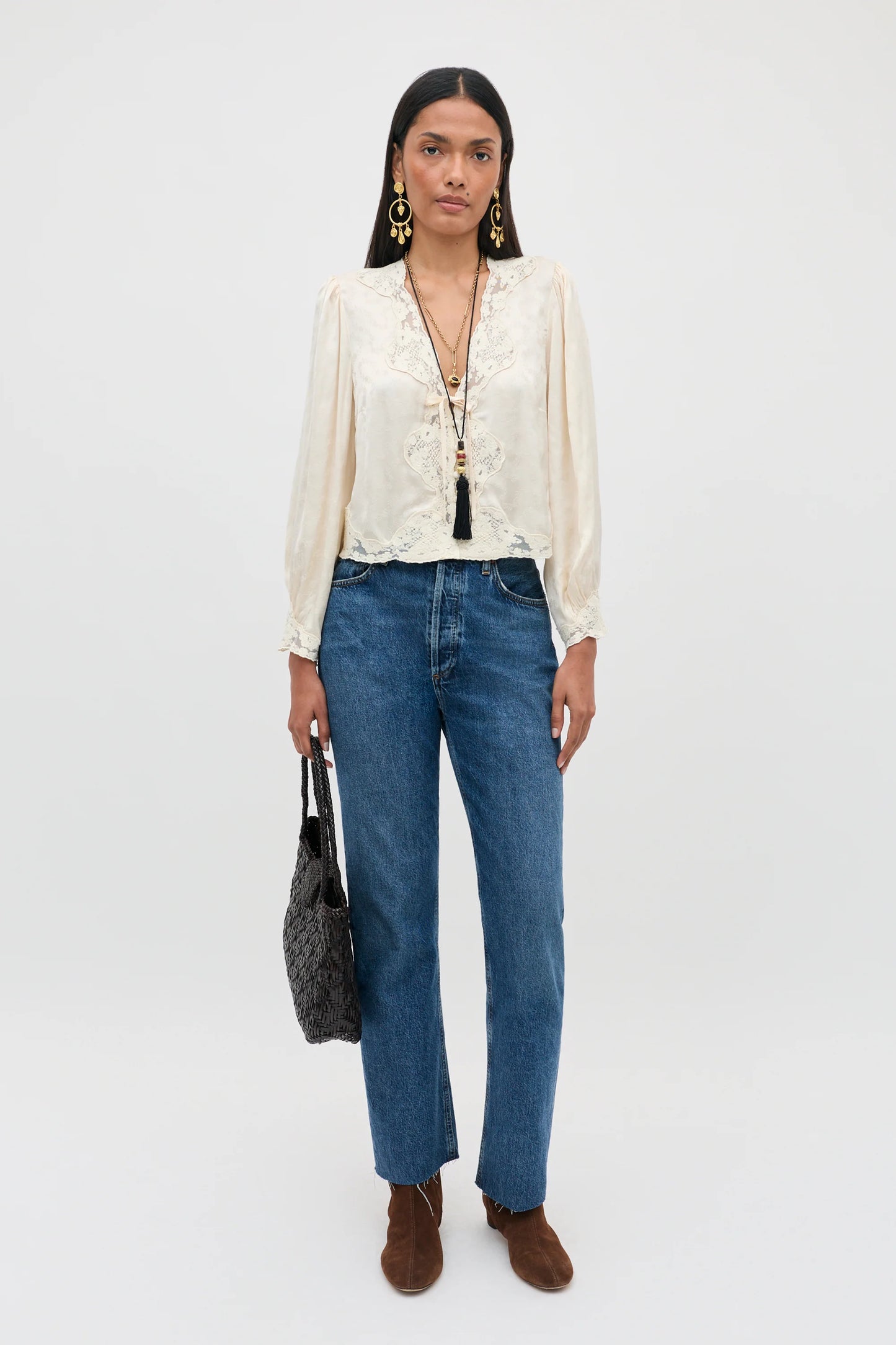 A model stands against a plain white studio background wearing a cream-colored lace-trimmed long-sleeve blouse, blue straight-leg jeans, brown suede boots, and holding a dark gray textured handbag.