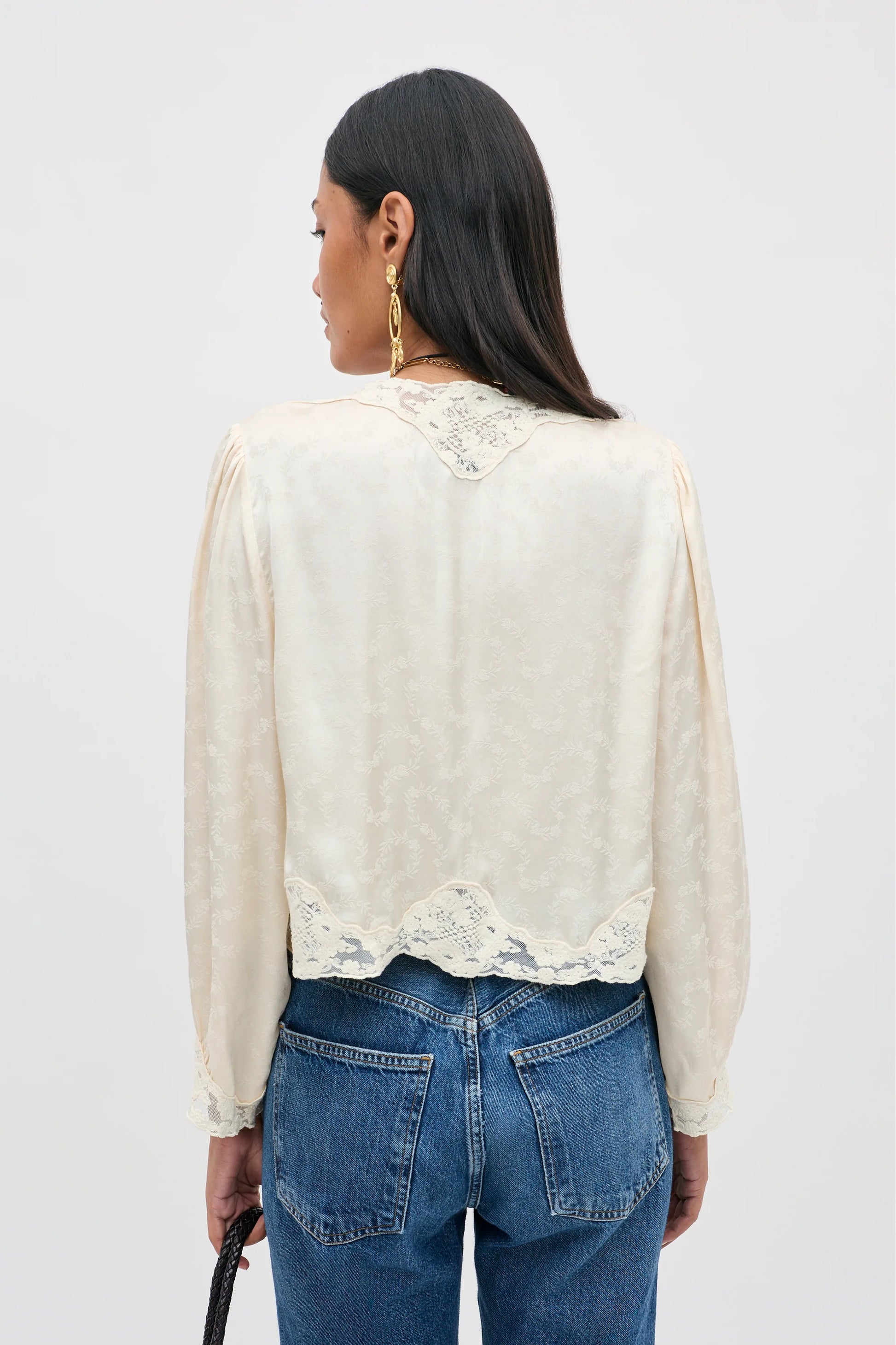A woman viewed from the back wearing a cream-colored long-sleeve blouse with lace trim at the neckline and hem, paired with blue jeans, against a plain light gray studio background.