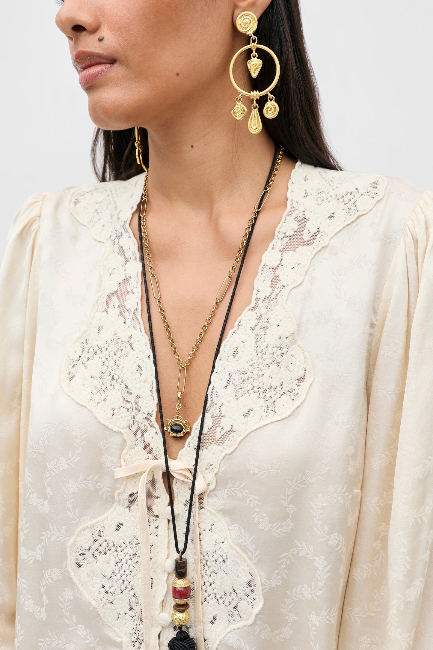 A close-up of a model wearing gold-toned jewelry, including a large circular dangle earring and layered necklaces, paired with a cream lace blouse.