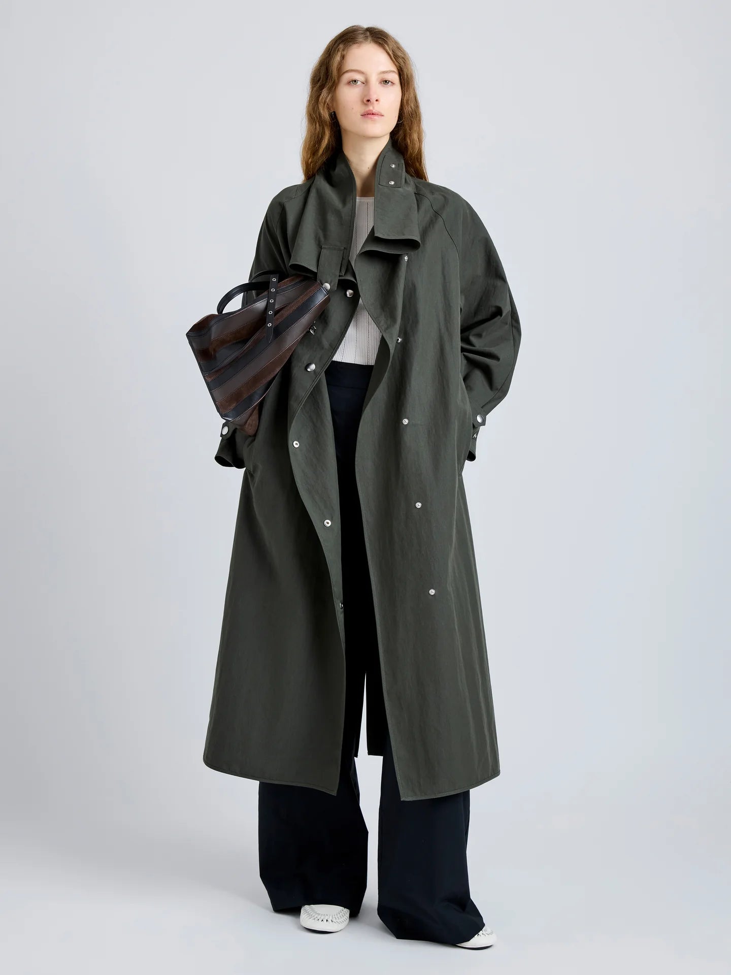 Woman wearing a long green coat holding a brown leather bag on a light gray background