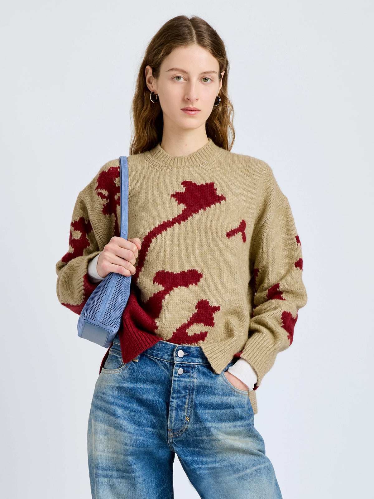 A woman modeling a beige knit sweater with a dark red abstract pattern, paired with blue jeans and holding a blue shoulder bag against a light gray studio background.