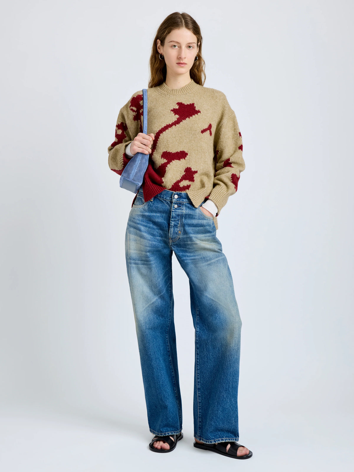 A woman stands against a light gray studio background wearing a beige knit sweater with a dark red abstract pattern, blue wide-leg jeans, black sandals, and carrying a blue shoulder bag.