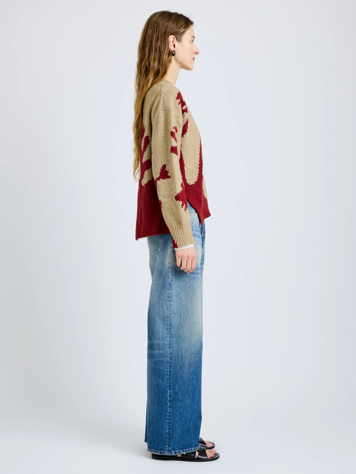 A woman stands in profile wearing a beige and burgundy knit sweater with a large abstract pattern, paired with wide-leg blue jeans and black sandals, against a light gray studio background.