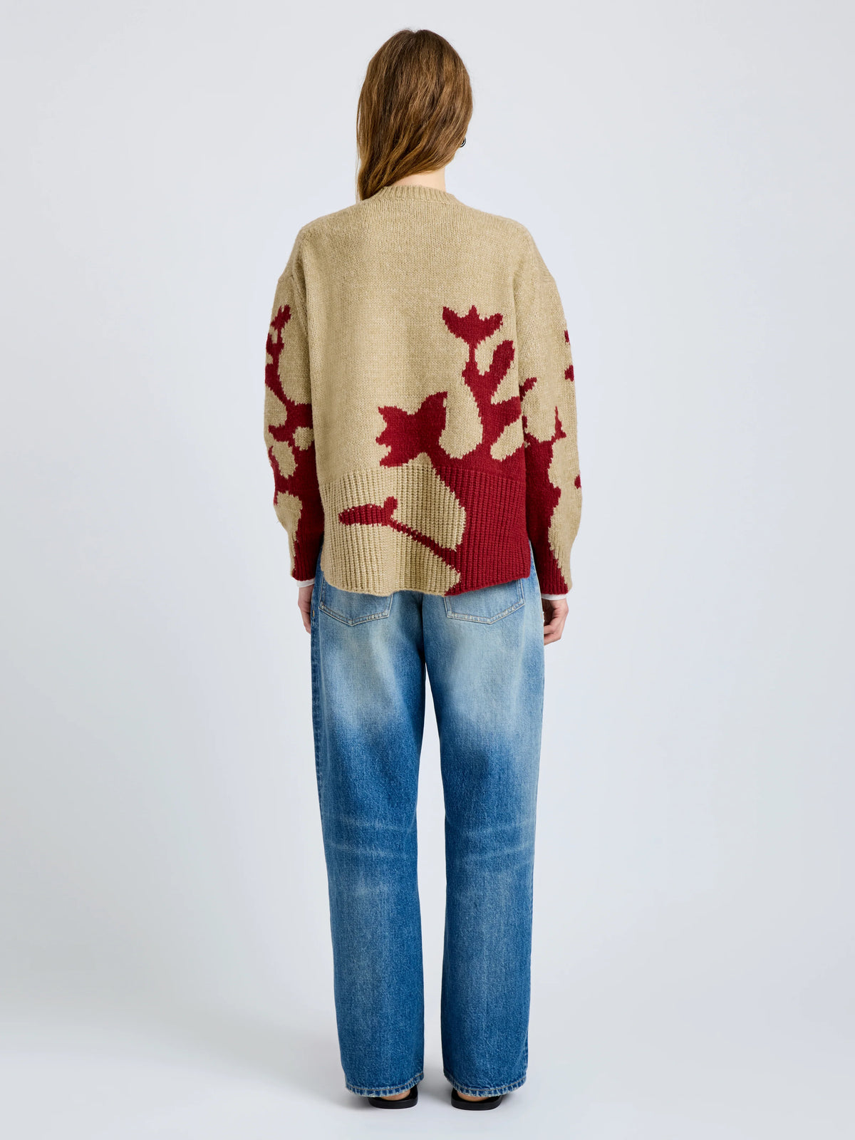 Person wearing a beige sweater with red abstract design and blue jeans on a white background