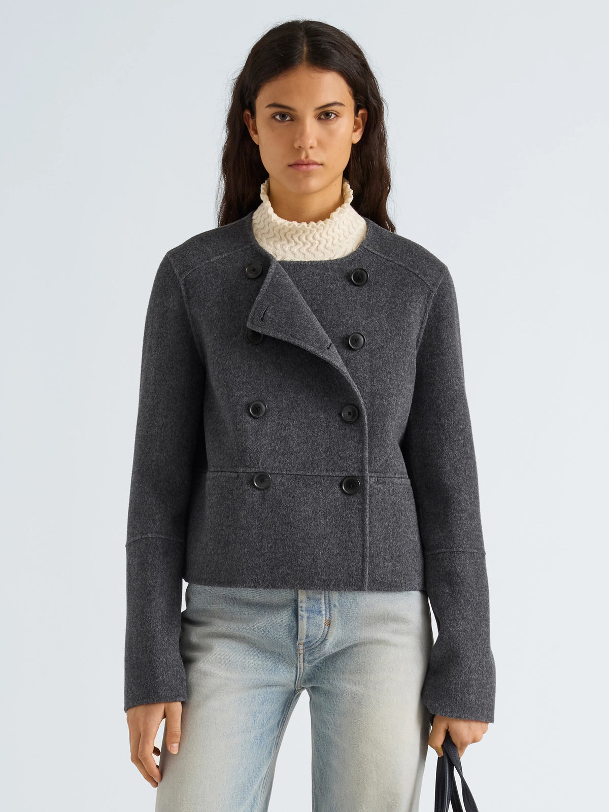 A woman modeling a dark gray double-breasted wool-blend jacket with an asymmetrical collar, worn over a cream knit turtleneck and light-wash jeans, against a light gray studio background.