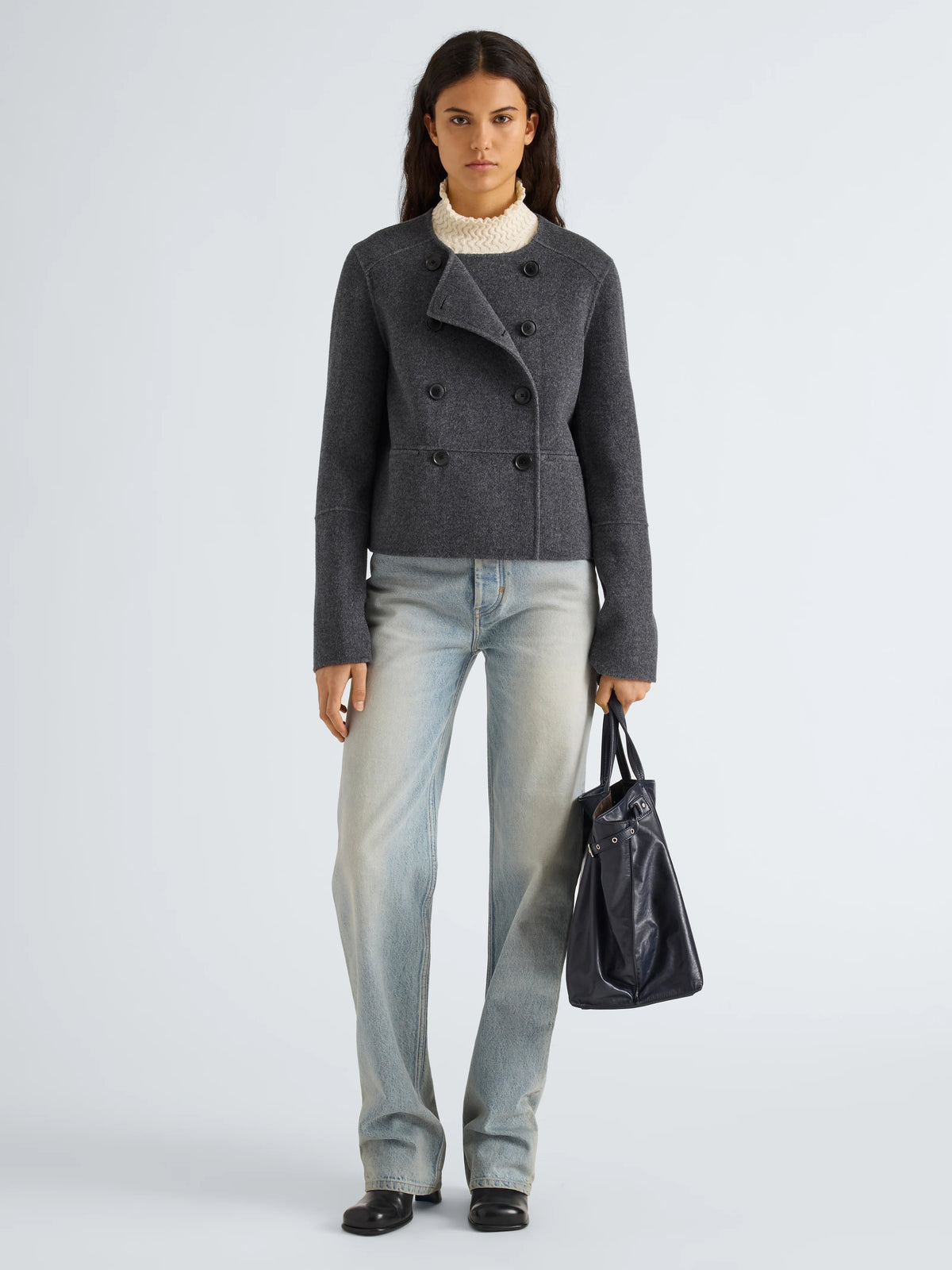 A model stands against a light gray studio background wearing a dark gray double-breasted wool jacket, light-wash blue jeans, a cream ribbed turtleneck, and black shoes, while holding a black leather tote bag.