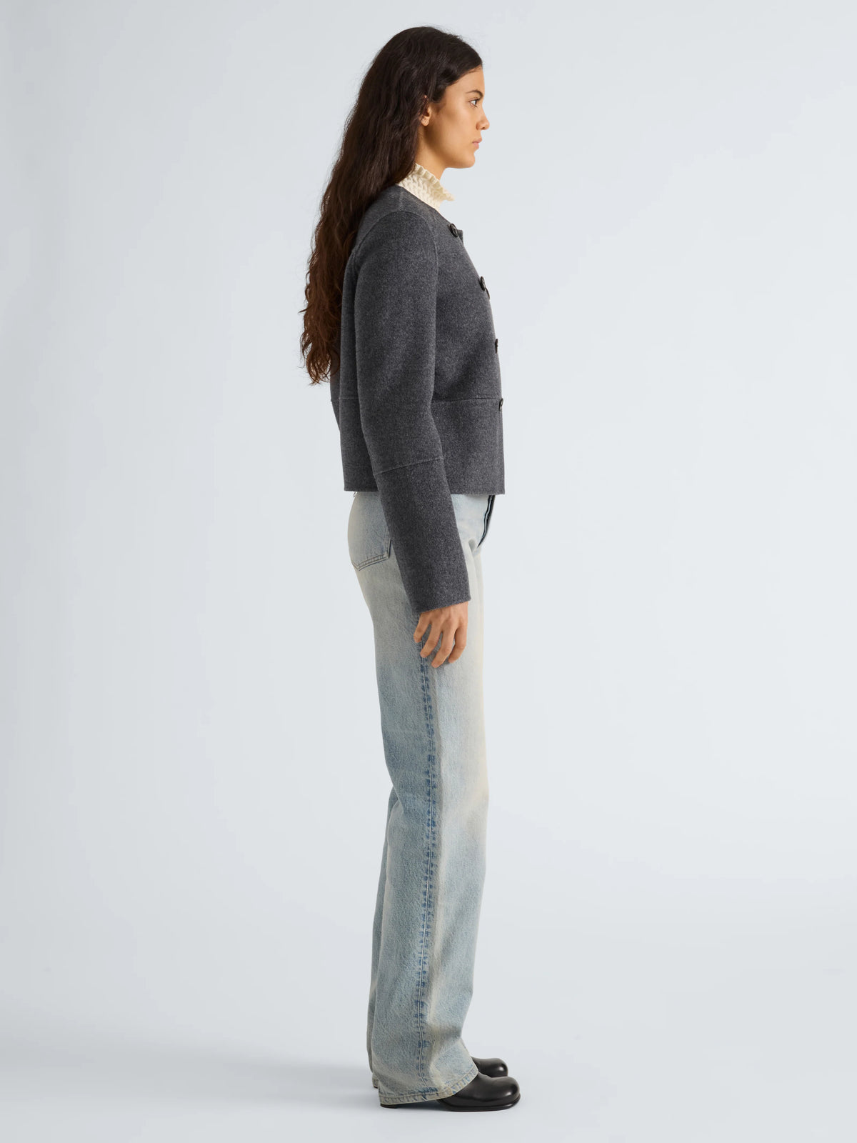 A woman shown in profile wearing a dark gray cropped knit cardigan, light-wash blue jeans, and black shoes against a plain light gray studio background.