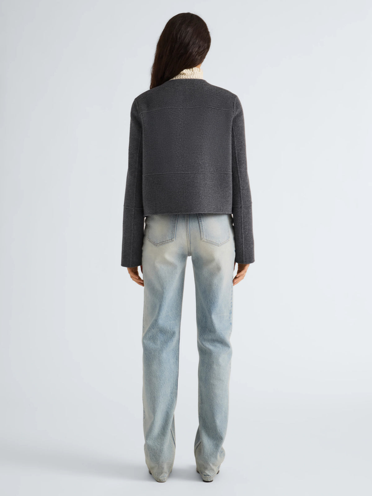A model shown from the back wearing a dark gray cropped knit cardigan and light-wash blue jeans against a light gray studio background.