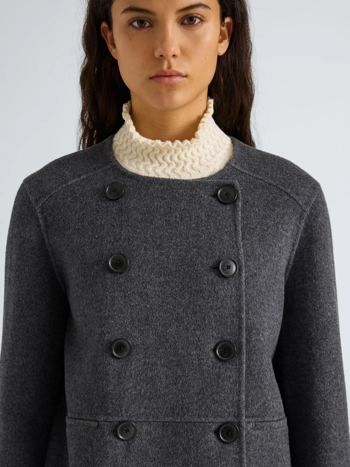 A close-up of a model wearing a dark gray double-breasted wool coat over a cream-colored knit mock neck.