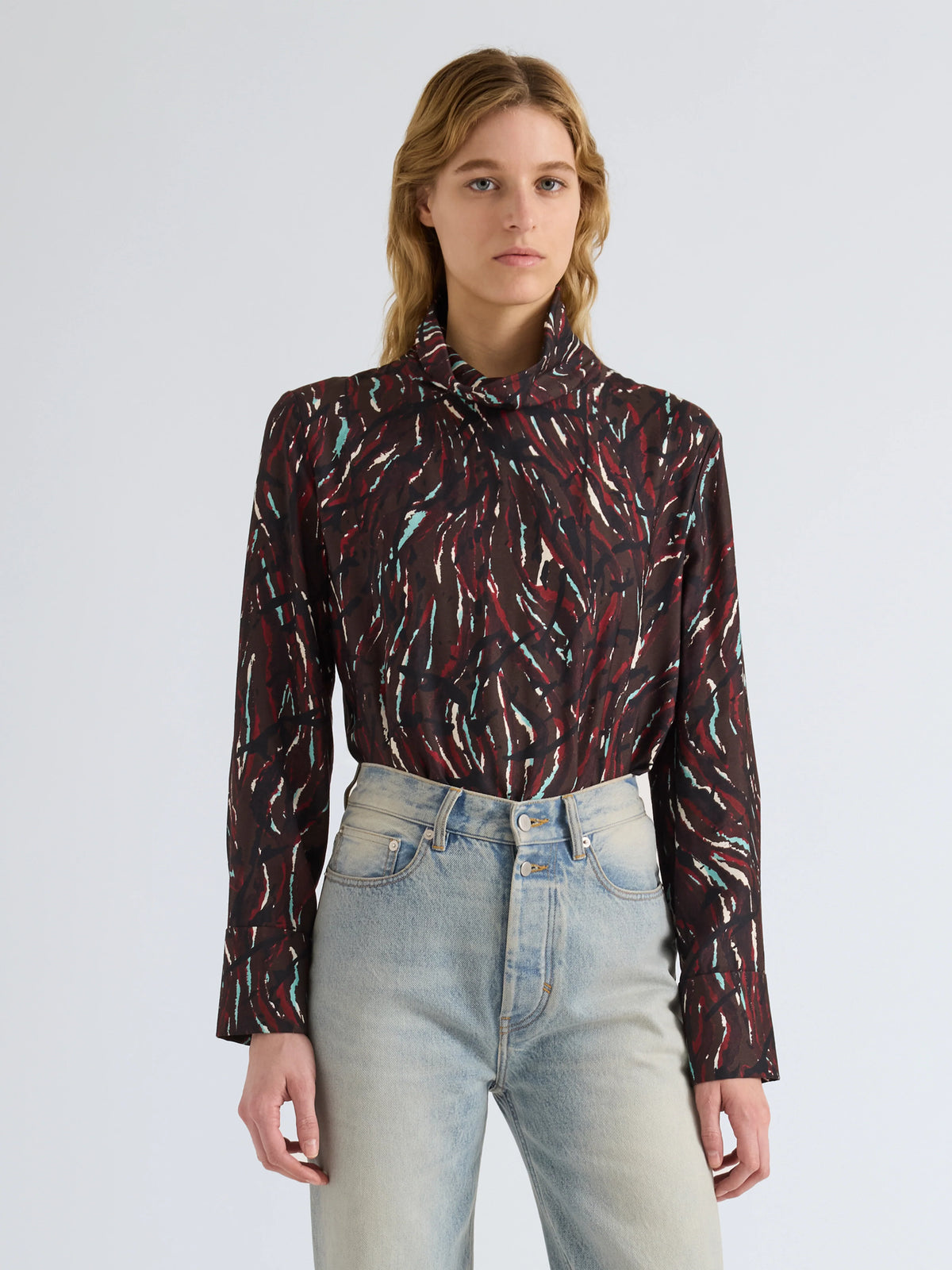 A woman modeling a long-sleeve high-neck blouse with an abstract brushstroke pattern in dark red, black, and light blue, paired with light-wash jeans against a light gray studio background.