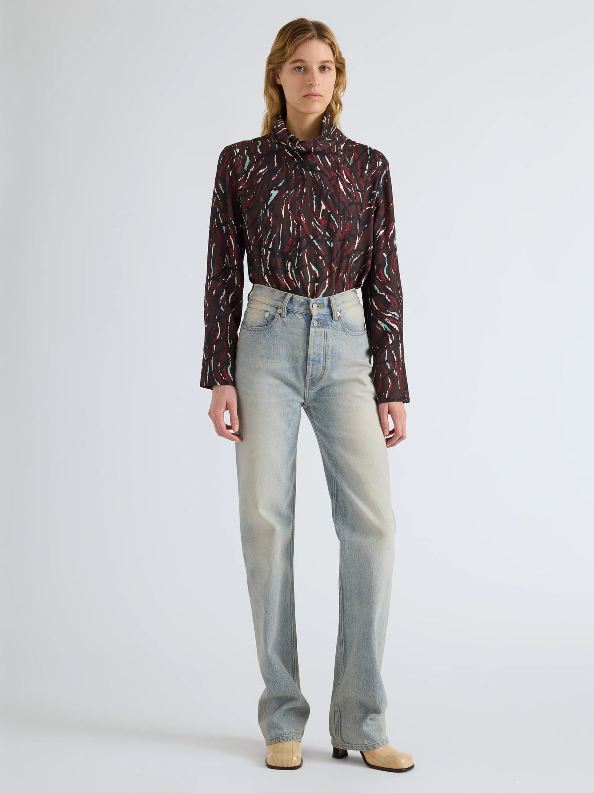 A model stands against a light gray studio background wearing a dark patterned long-sleeve blouse with a high neck and light-wash straight-leg jeans.