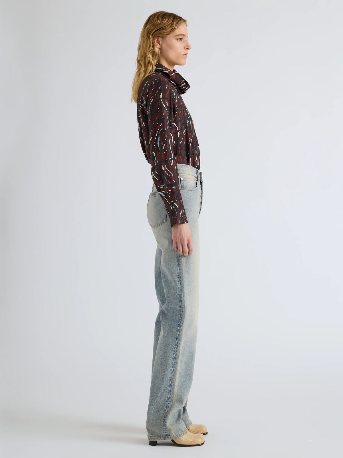 A woman stands in profile wearing a dark patterned long-sleeve shirt, light-wash straight-leg jeans, and beige shoes against a plain light gray studio background.