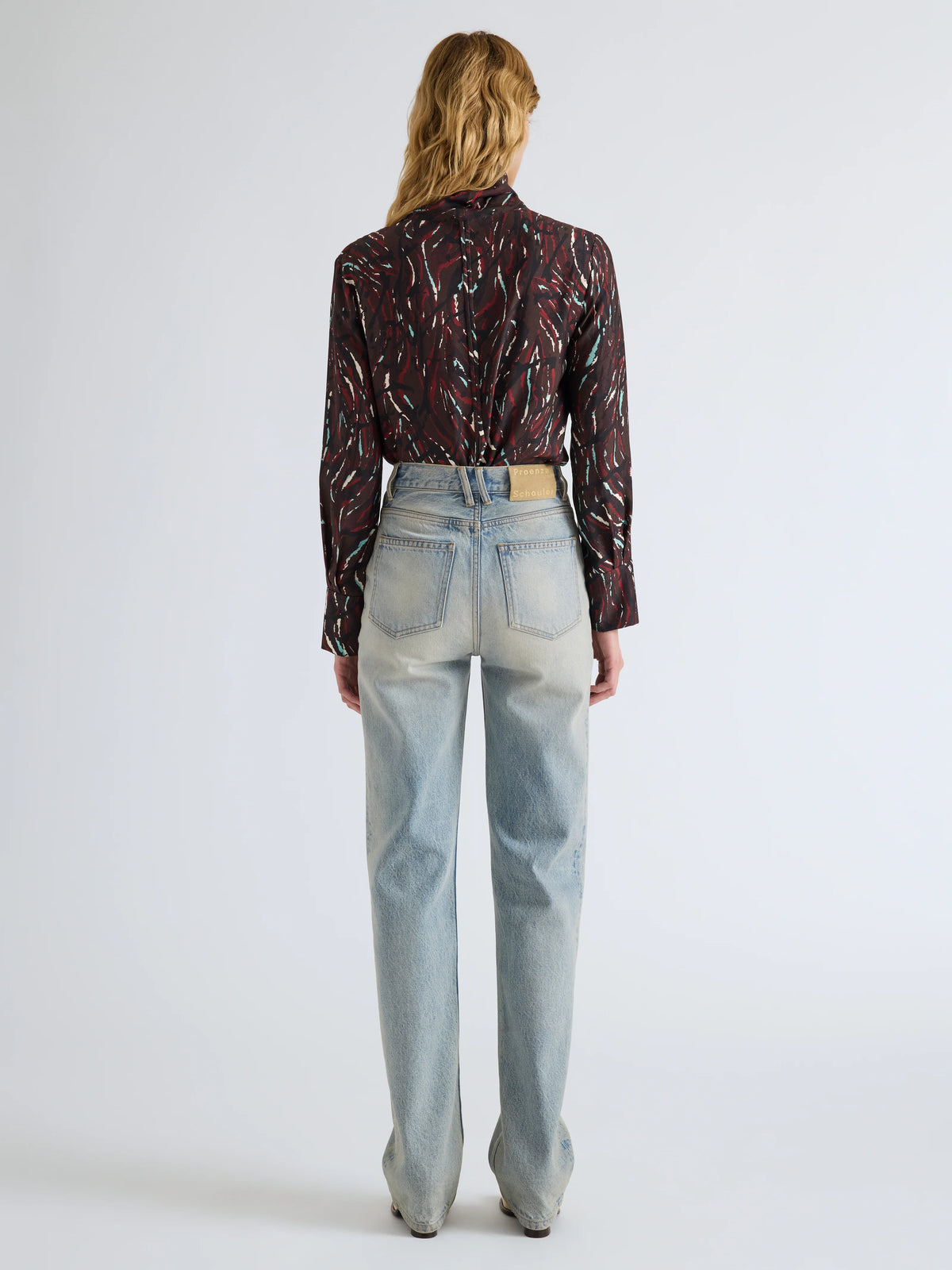 A woman viewed from behind wearing a dark patterned long-sleeve shirt and light-wash straight-leg jeans against a plain light gray studio background.