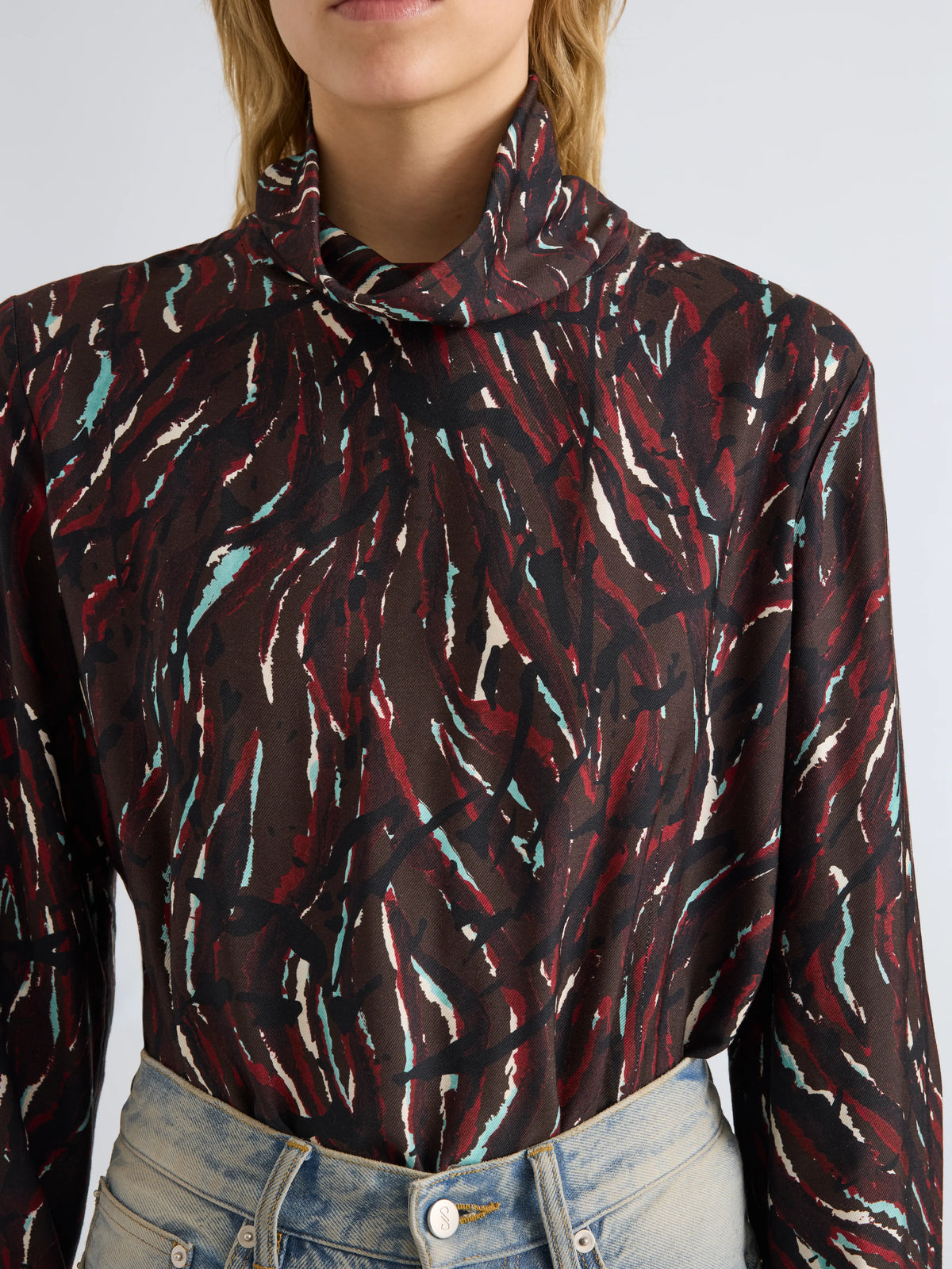 A close-up of a model wearing a long-sleeve mock neck blouse with an abstract brushstroke pattern in black, red, and white, paired with light-wash blue jeans.