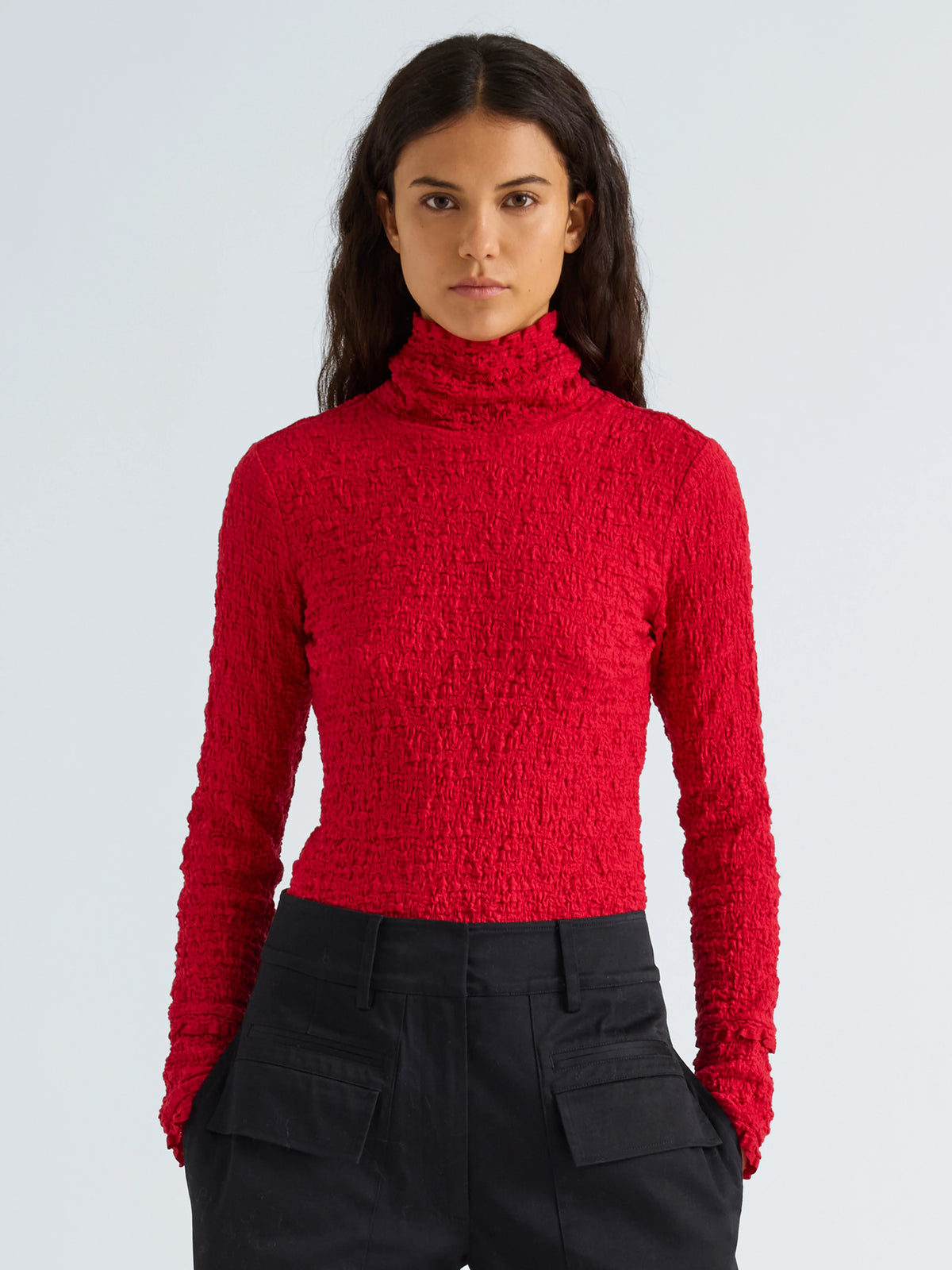 A woman modeling a textured, bright red long-sleeve mock neck top paired with black cargo pants against a light gray studio background.