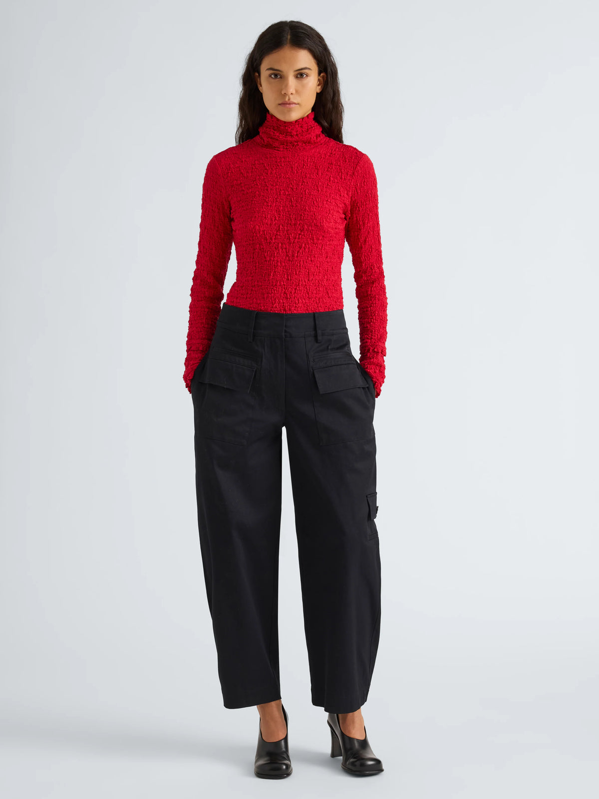 A model stands against a light gray studio background wearing a bright red textured knit turtleneck sweater and black wide-leg cropped trousers with black shoes.