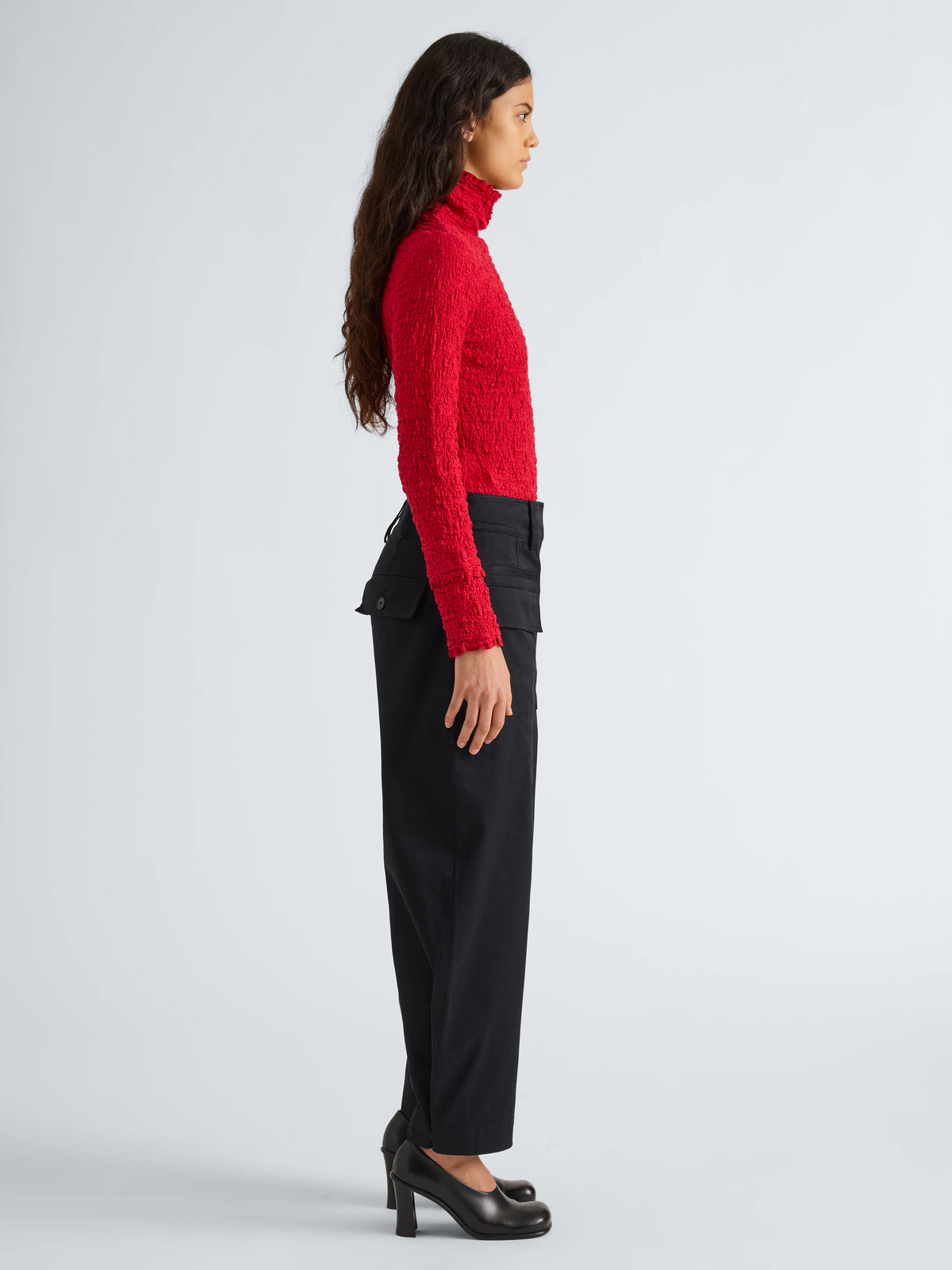 A woman stands in profile wearing a bright red textured turtleneck sweater, black wide-leg trousers, and black high heels against a light gray studio background.