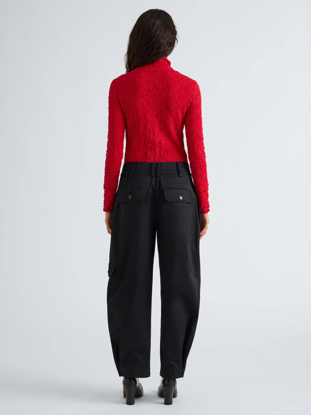 A model viewed from the back wearing a bright red textured knit mock-neck sweater and black wide-leg trousers, set against a plain light gray studio background.