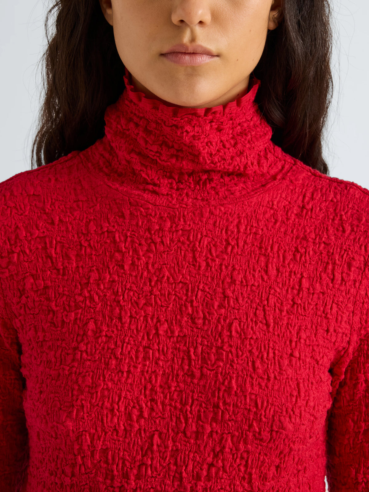 A close-up of a model wearing a bright red textured knit turtleneck sweater against a light gray background.