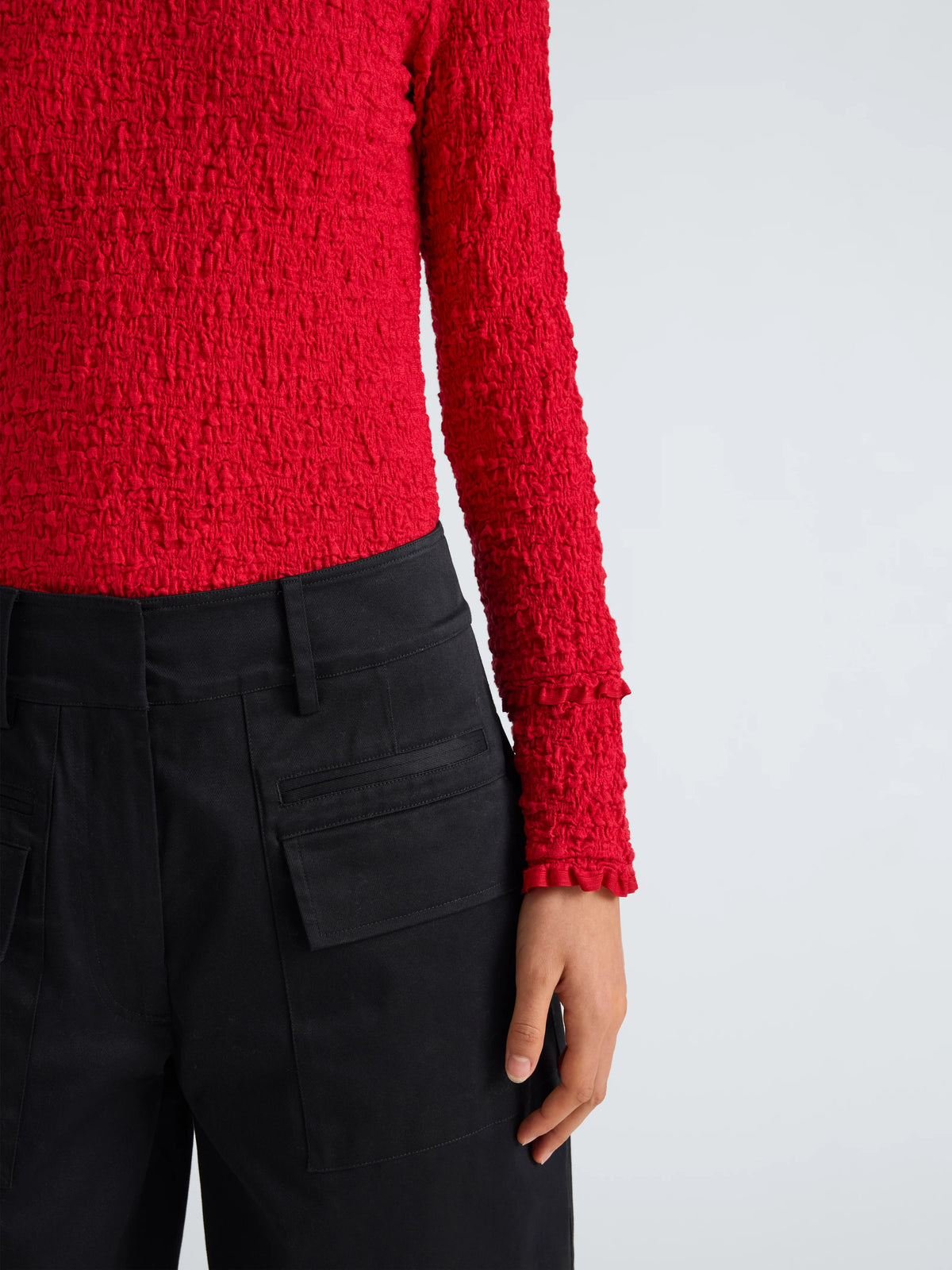 A close-up of a person wearing a bright red textured long-sleeve top and black trousers, shown from the chest down against a light gray studio background.
