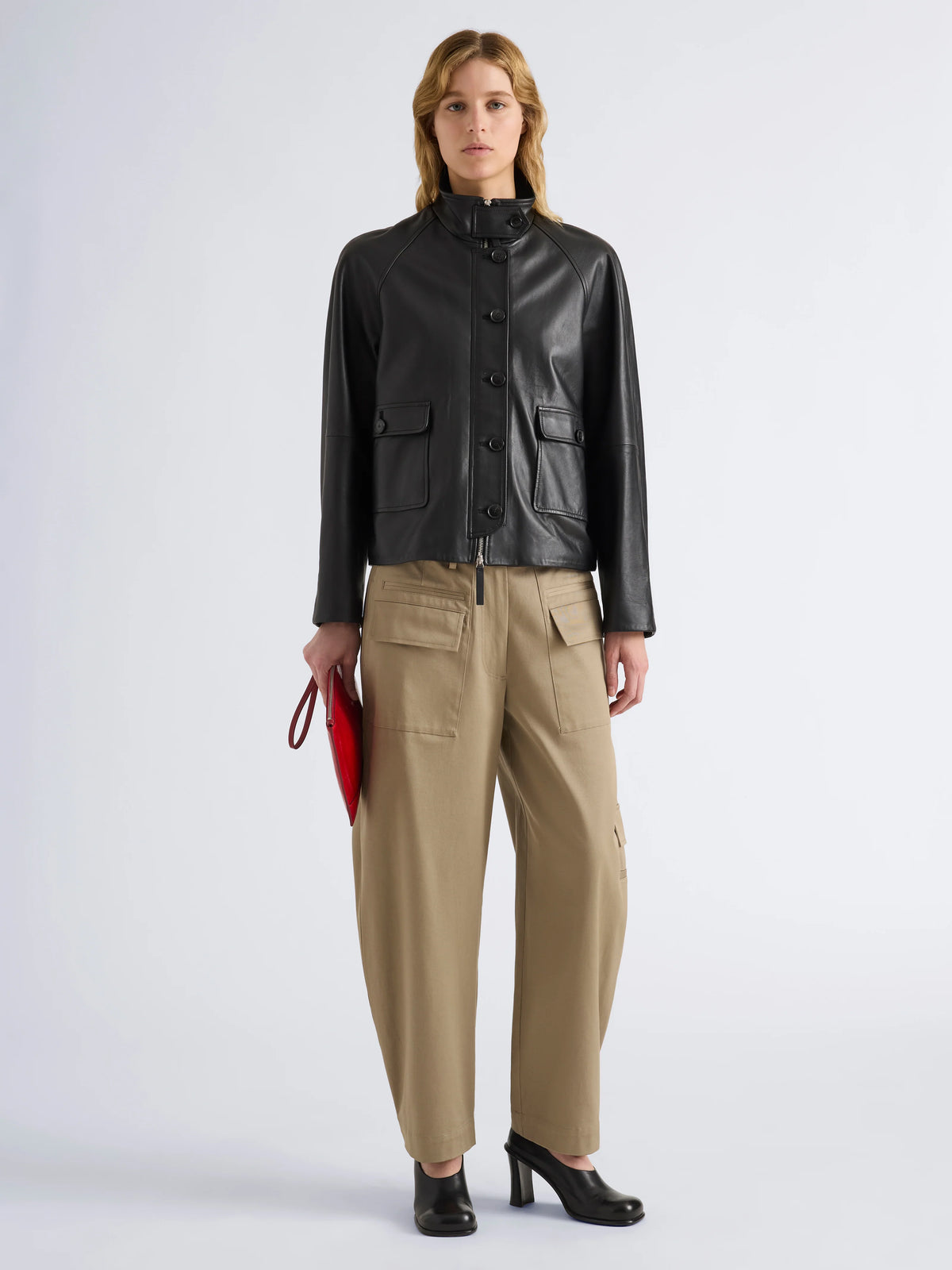A model stands against a light gray studio background wearing a black leather jacket with a high collar and front buttons, paired with beige wide-leg trousers and black shoes. She holds a small red clutch in one hand.