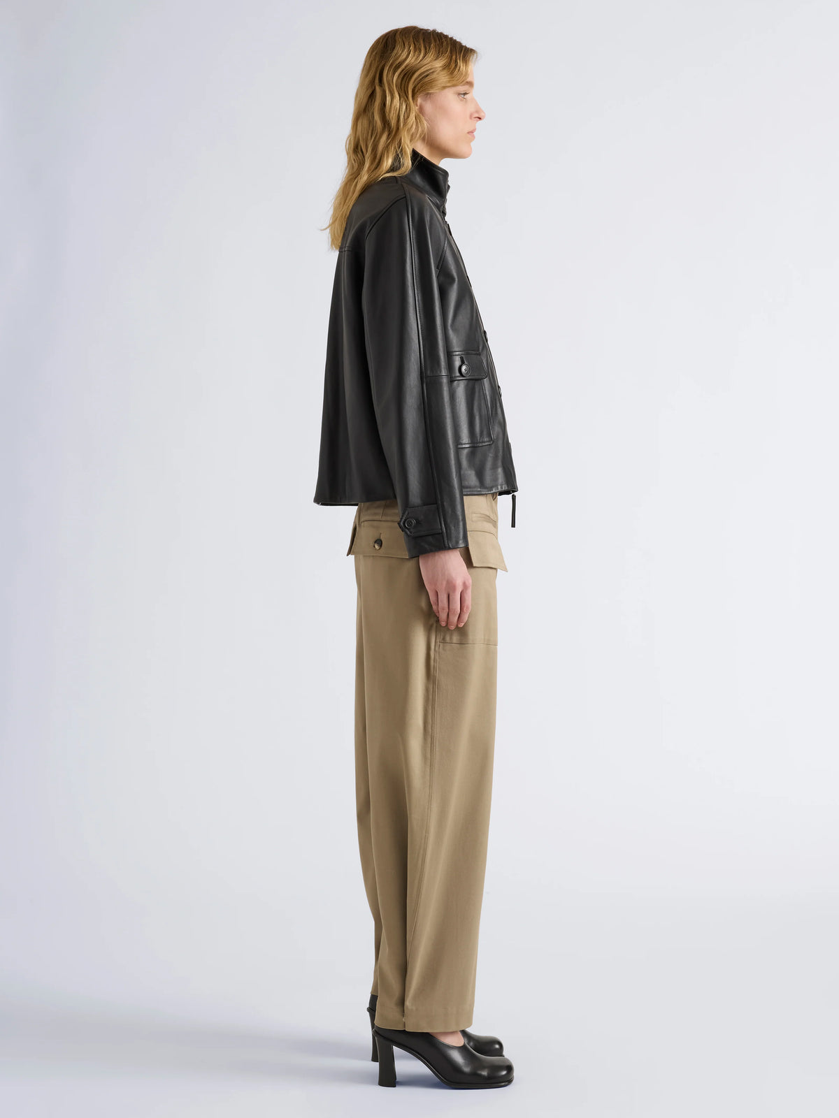 A model shown in profile wearing a black leather jacket, beige wide-leg trousers, and black high heels against a light gray studio background.