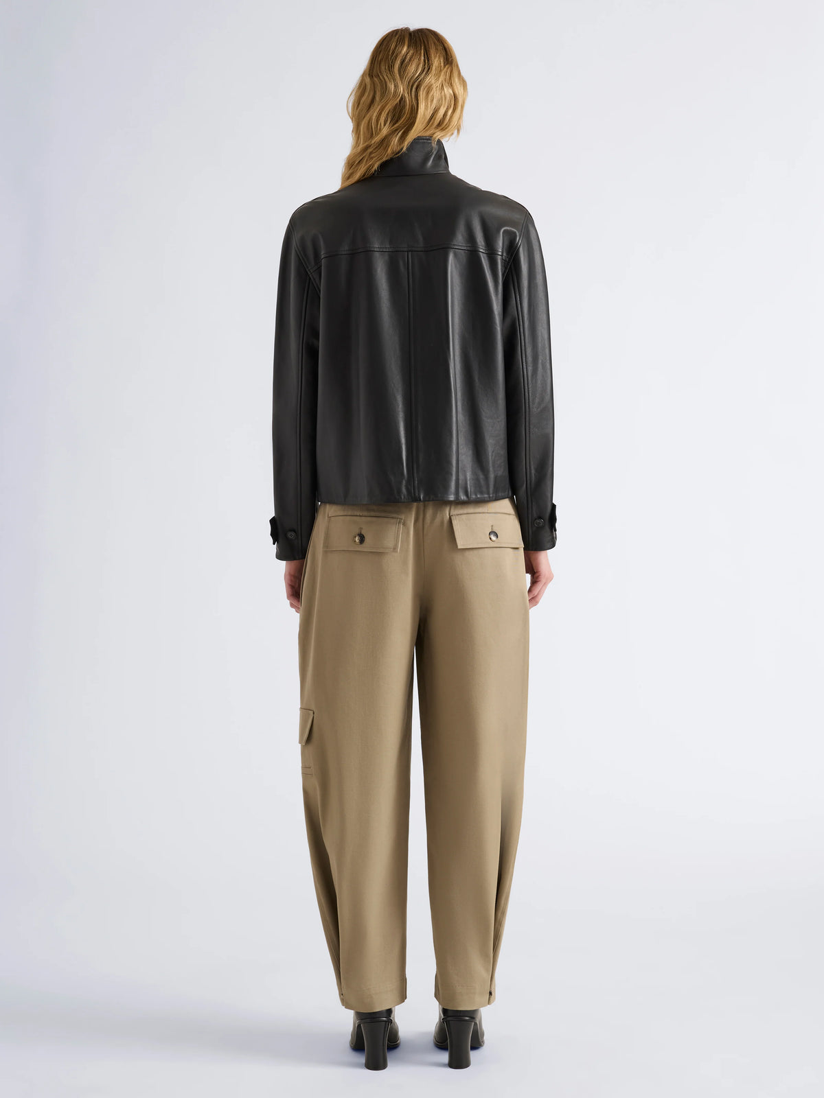 A model viewed from the back wearing a black leather jacket, beige wide-leg cargo pants, and black high heels against a light gray studio background.