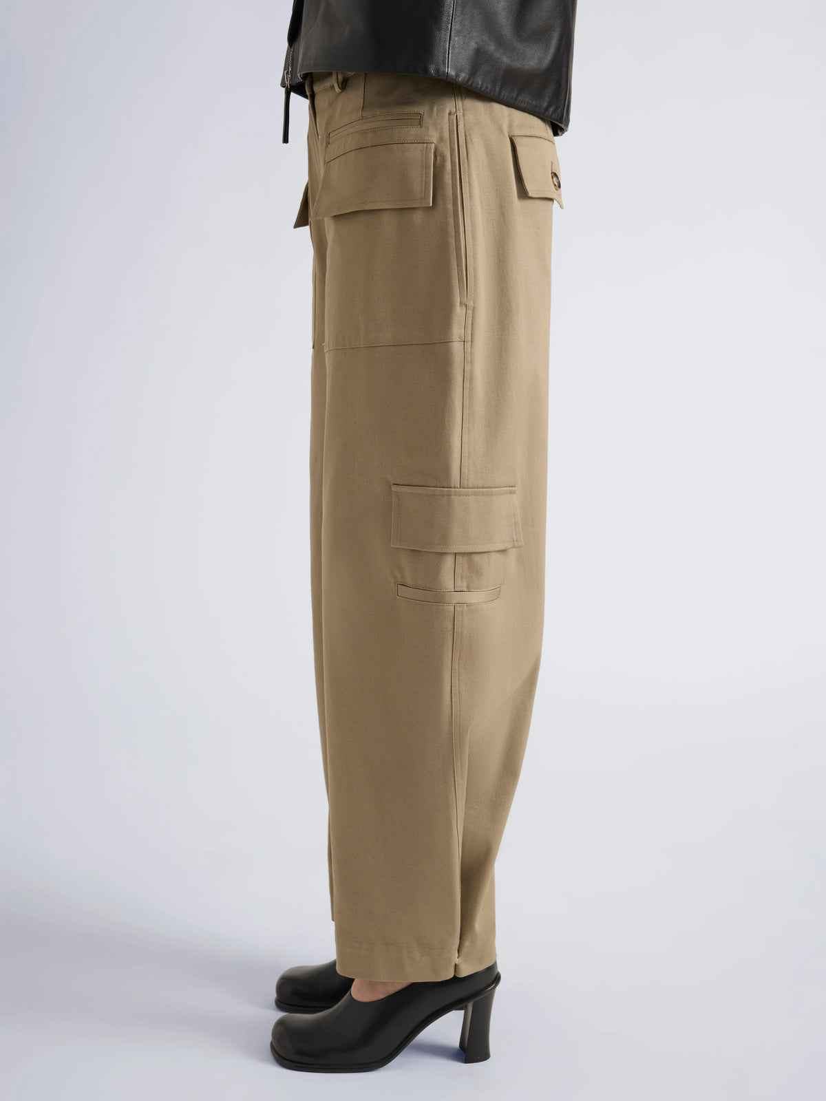 A model shown in profile wearing a beige wide-leg trousers, and black high heels against a light gray studio background.
