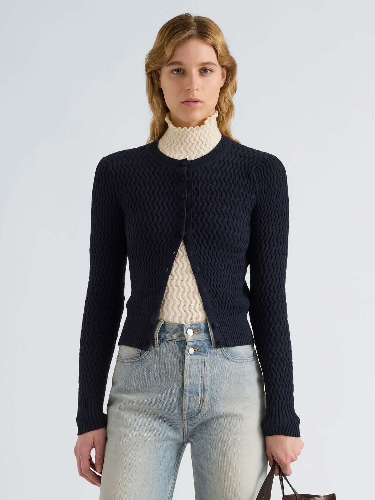 A woman modeling a dark navy blue textured knit cardigan over a cream ribbed turtleneck sweater and light-wash blue jeans, against a plain light gray studio background.