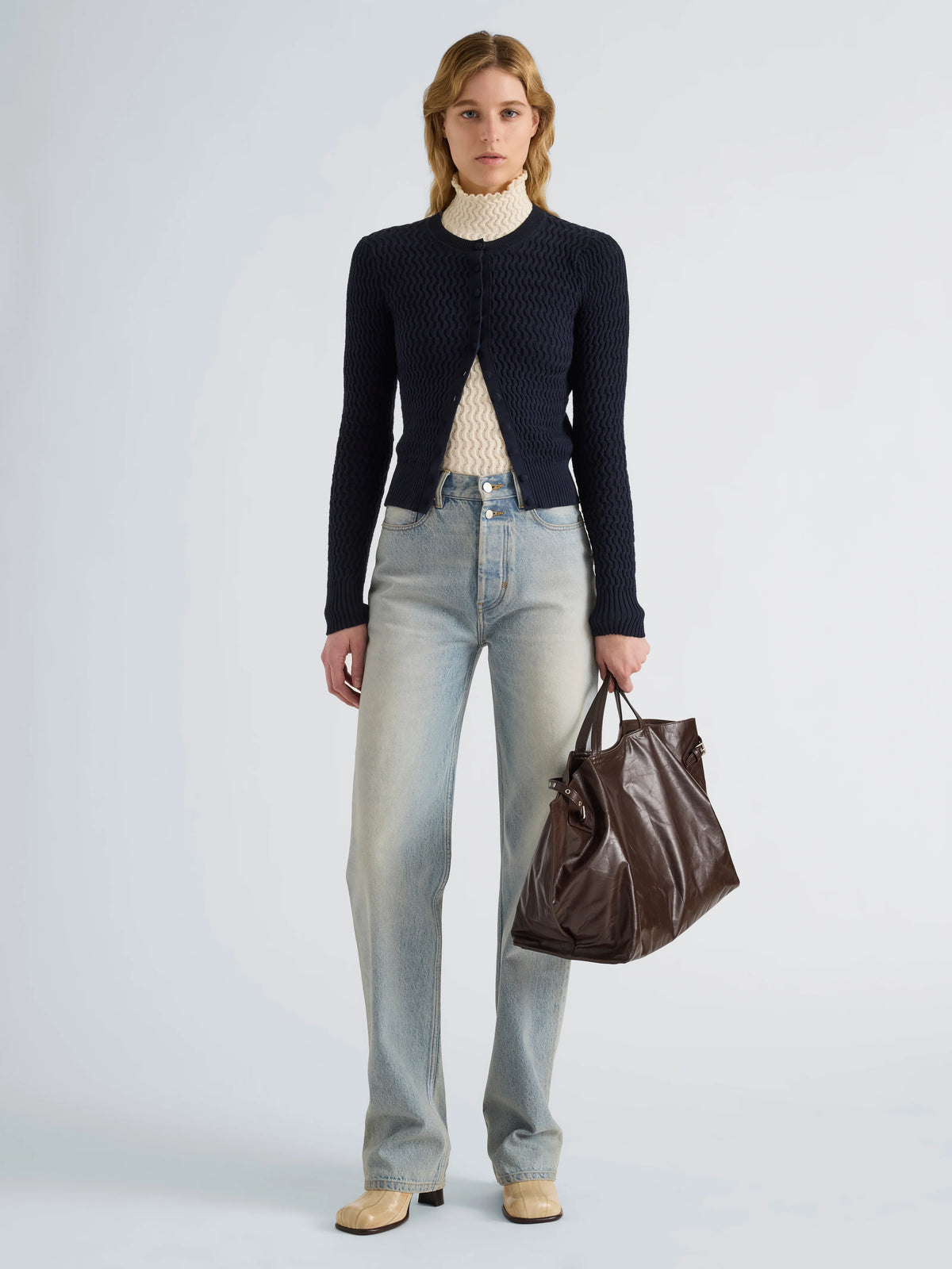 A model stands against a light gray studio background wearing a dark navy textured cardigan over a cream turtleneck, light-wash blue jeans, beige shoes, and holding a dark brown leather tote bag.