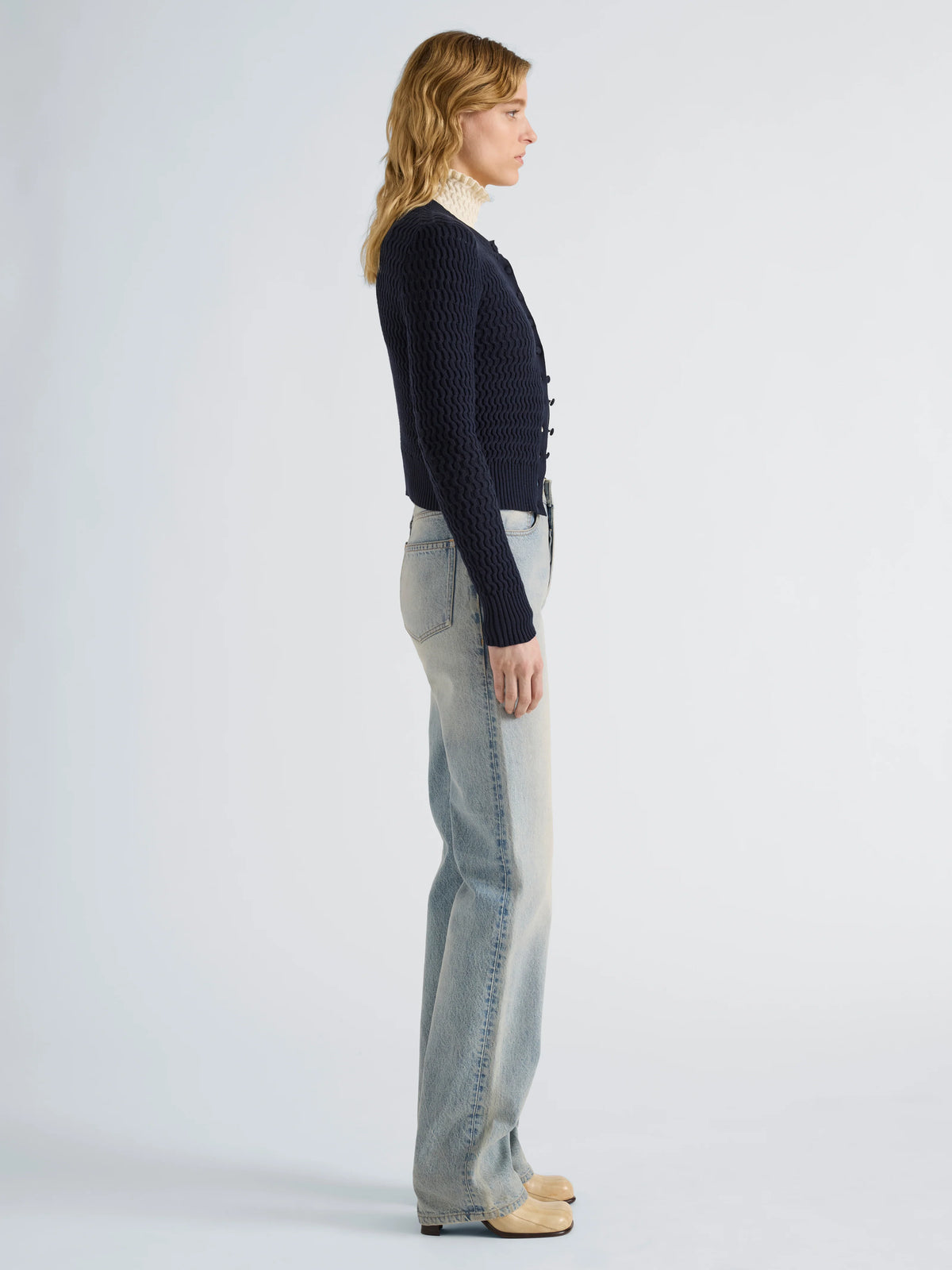 A woman stands in profile wearing a dark navy textured knit cardigan, light-wash wide-leg jeans, and tan shoes against a plain light gray studio background.
