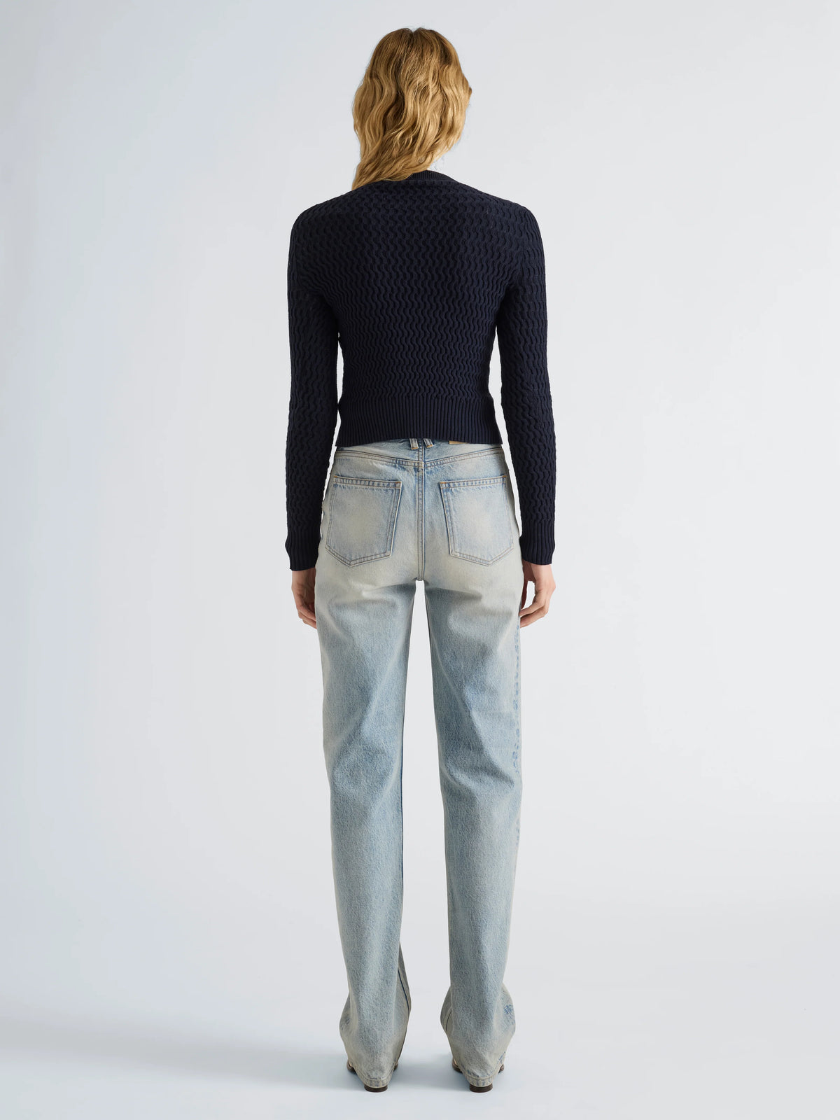 A woman viewed from the back wearing a dark navy textured knit long-sleeve top and light-wash blue jeans, standing against a plain light gray studio background.