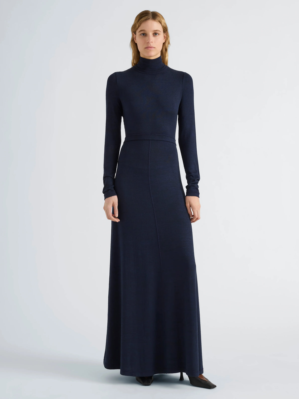A woman modeling a long-sleeve, high-neck, floor-length navy blue knit dress against a light gray studio background.