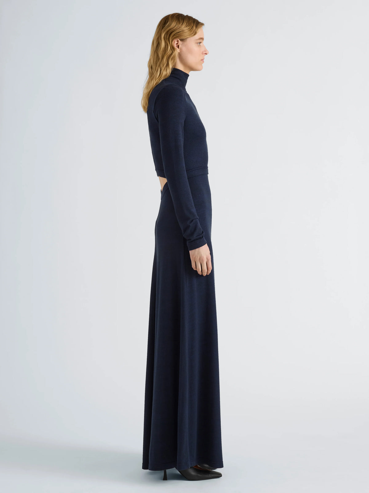 A woman in profile wearing a long-sleeve, high-neck, floor-length navy dress against a light gray studio background.