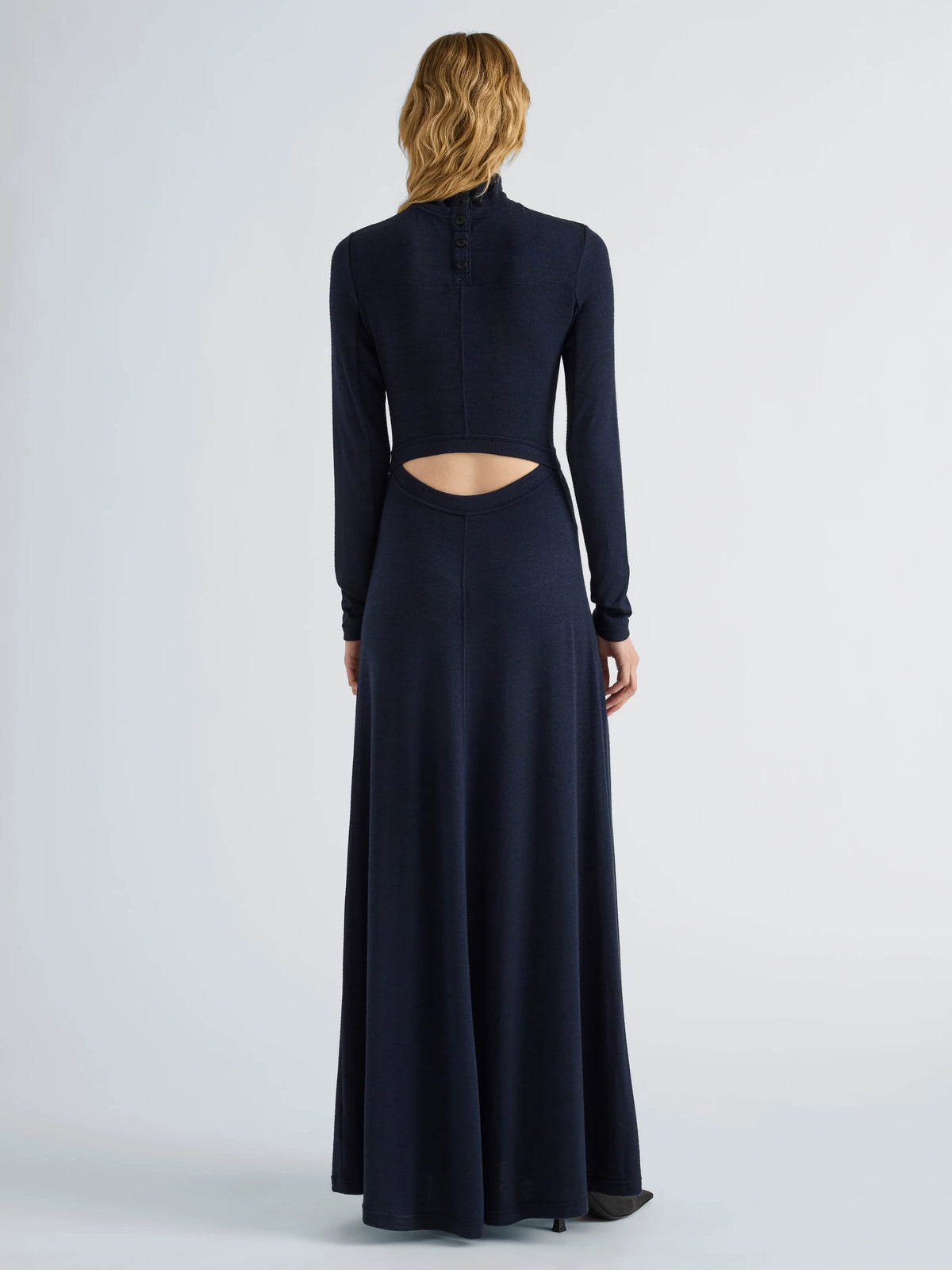 A woman viewed from the back wearing a long-sleeve navy maxi dress with a high mock neck and a cutout at the waist, photographed against a light gray studio background.