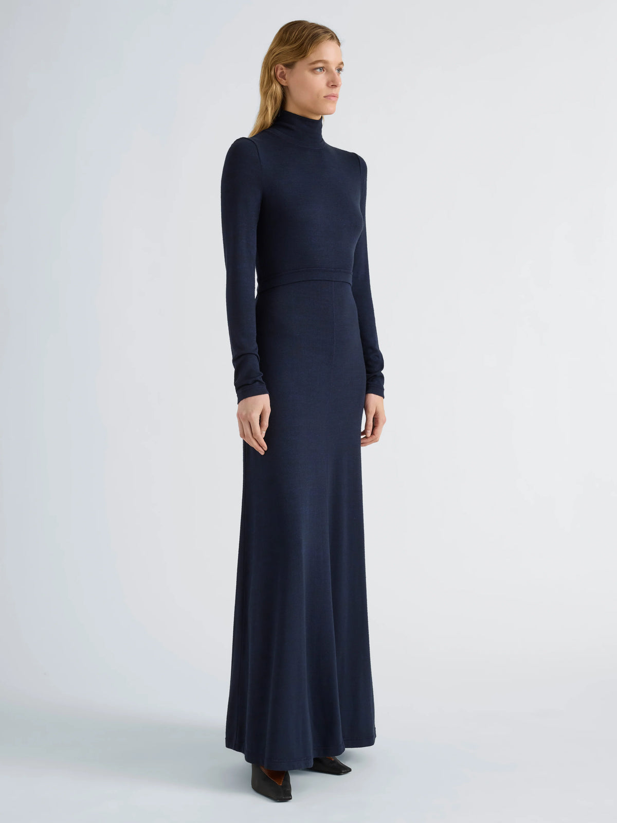 A woman modeling a long-sleeve, high-neck, floor-length navy blue knit dress against a light gray studio background.