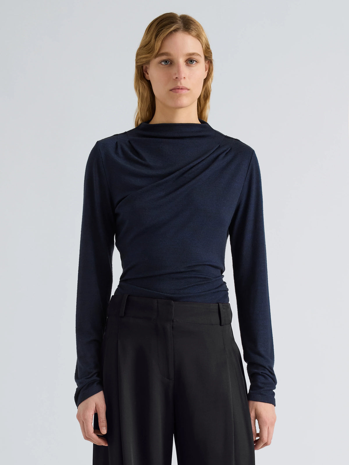 A model stands against a light gray studio background wearing a dark navy long-sleeve draped top