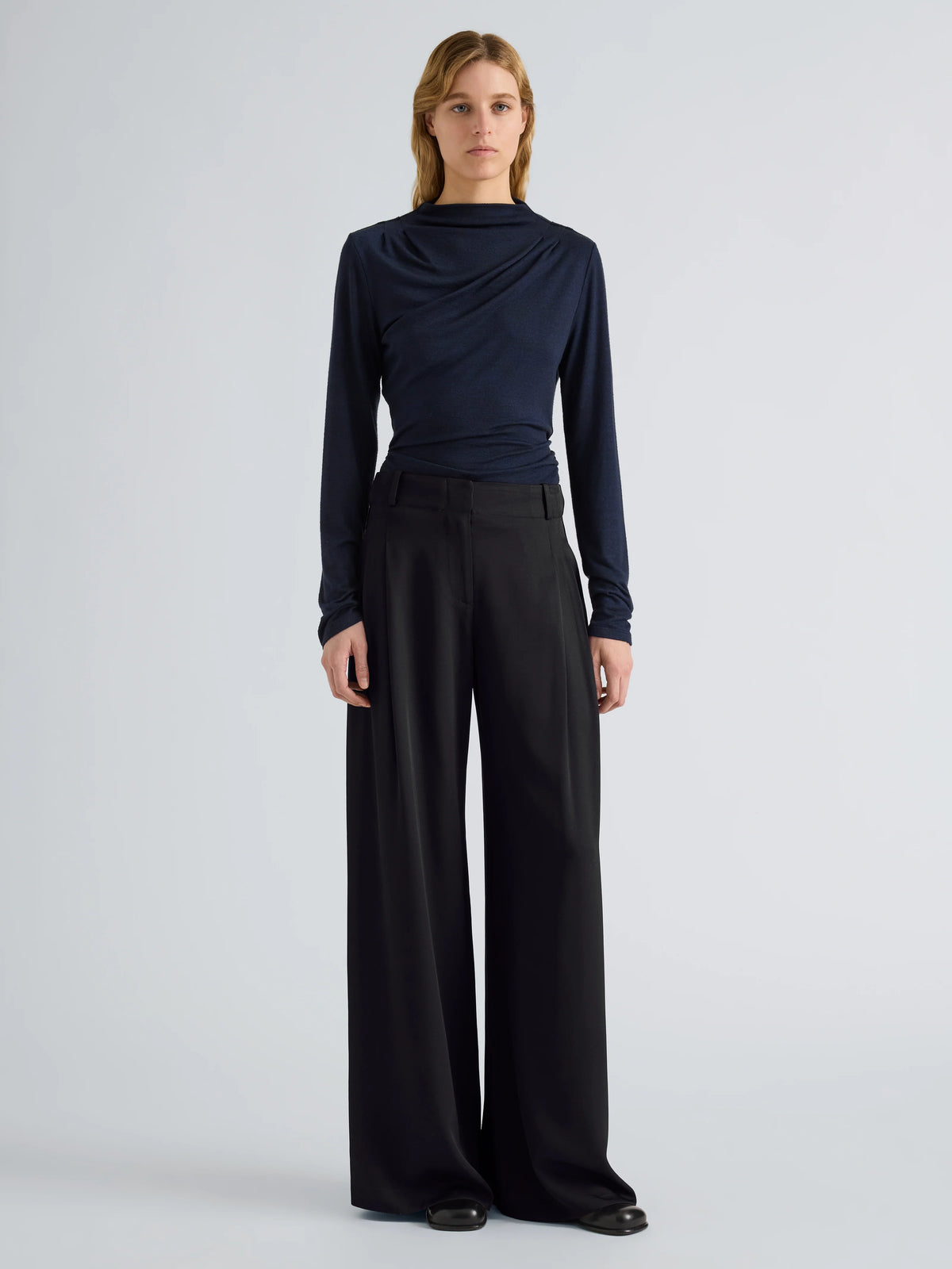 A model stands against a light gray studio background wearing a dark navy long-sleeve draped top and black wide-leg trousers.